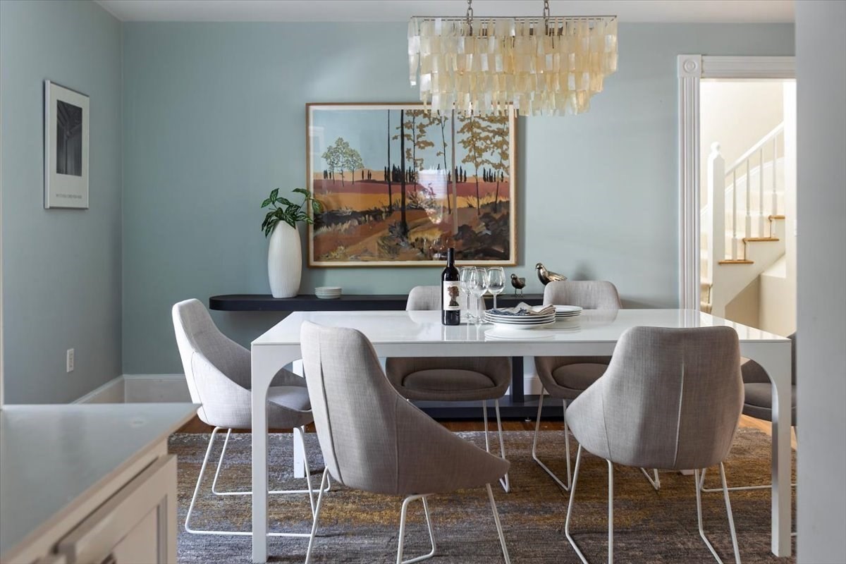 Dining room with a white table, upholstered chairs, chandelier, and staircase beyond the doorway.