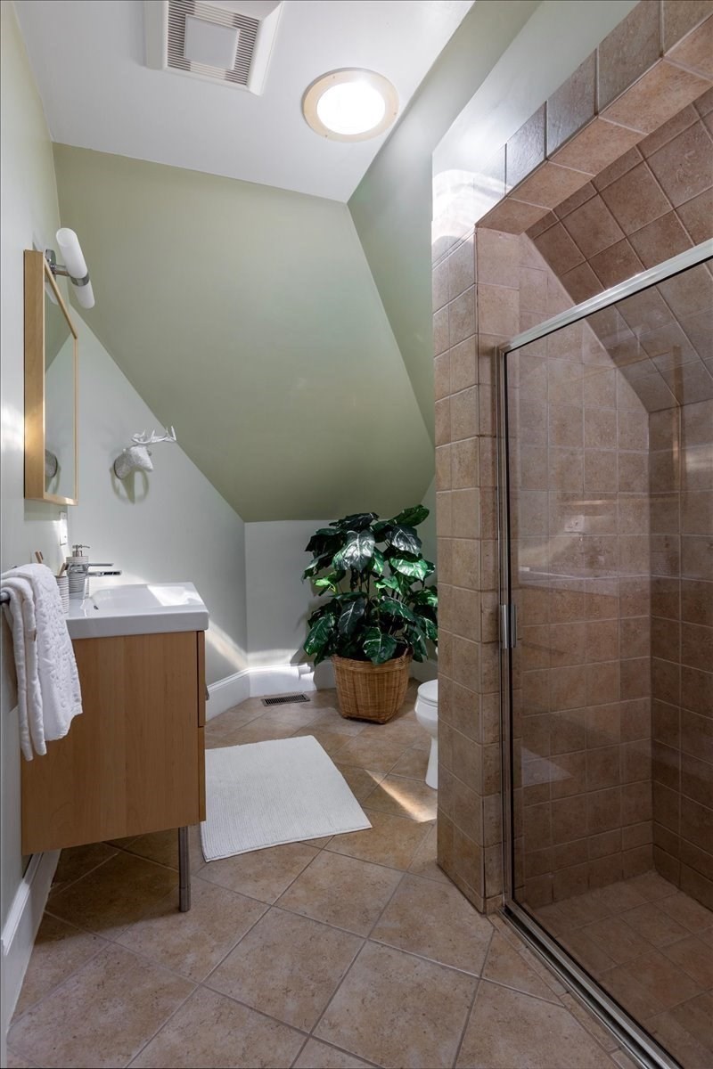 Bathroom with a glass-enclosed tan-tile shower, wood vanity, and sloped ceiling.