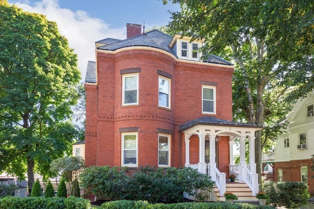 Red brick home with a curved bay and white-columned front porch amid mature trees.
