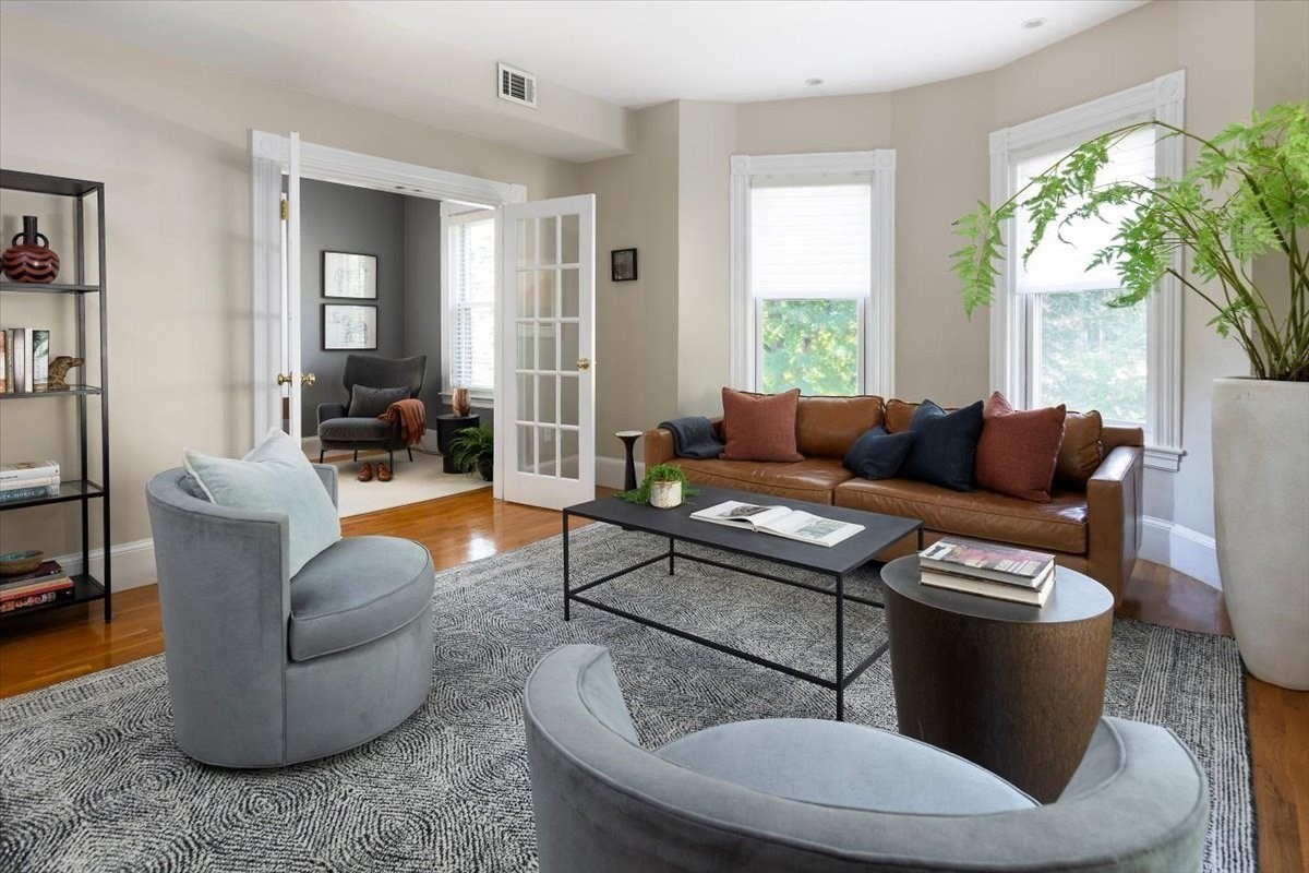 Staged living room with a leather sofa, curved gray chairs, tall windows, and French doors to a sitting room.