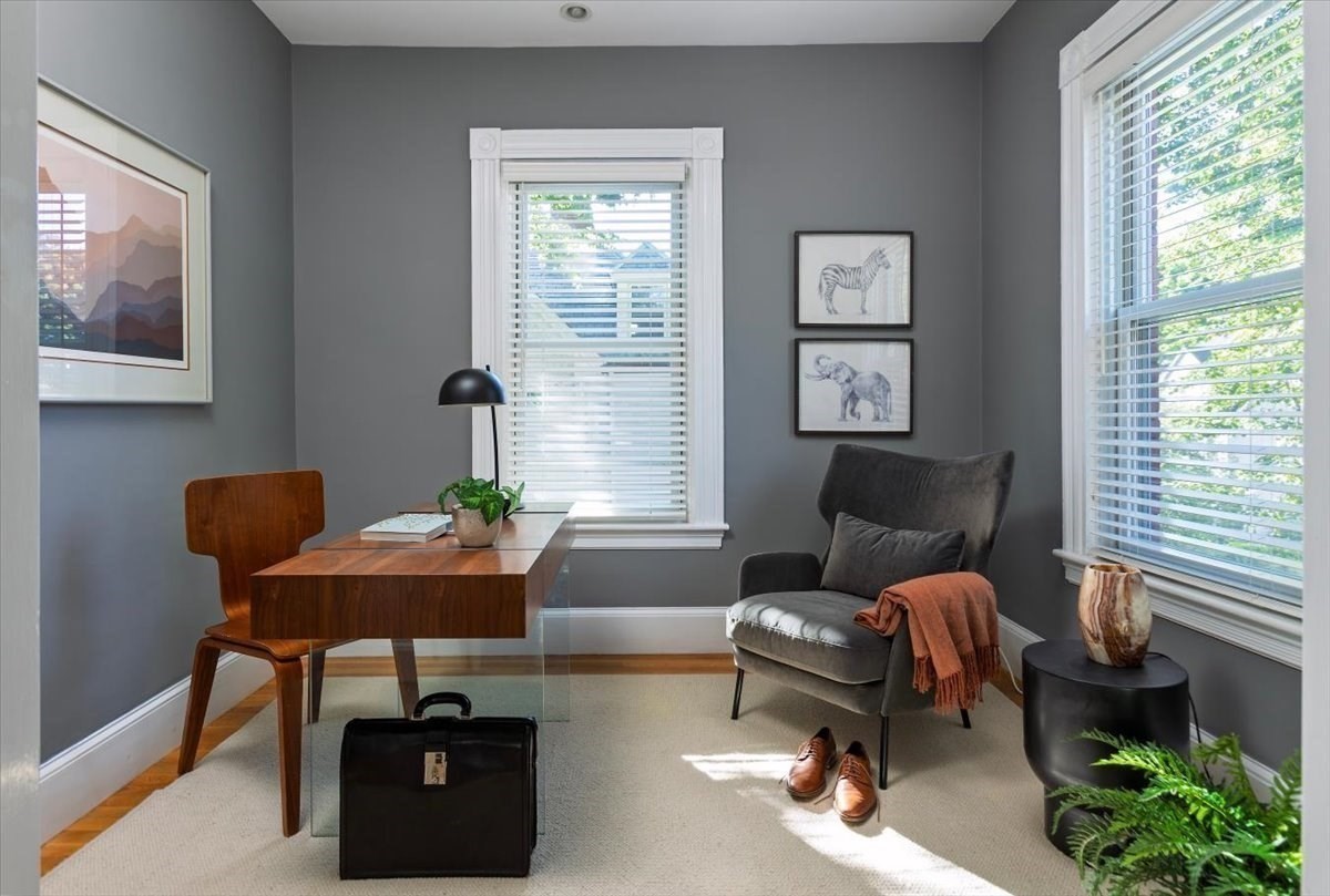 Furnished home office with a wooden desk, gray reading chair, and bright windows.