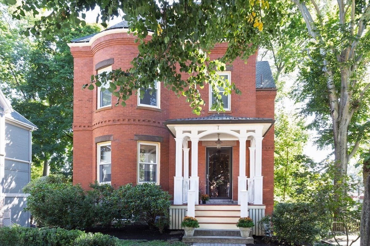 Red brick home with a rounded bay window and white-columned front porch beneath mature trees.