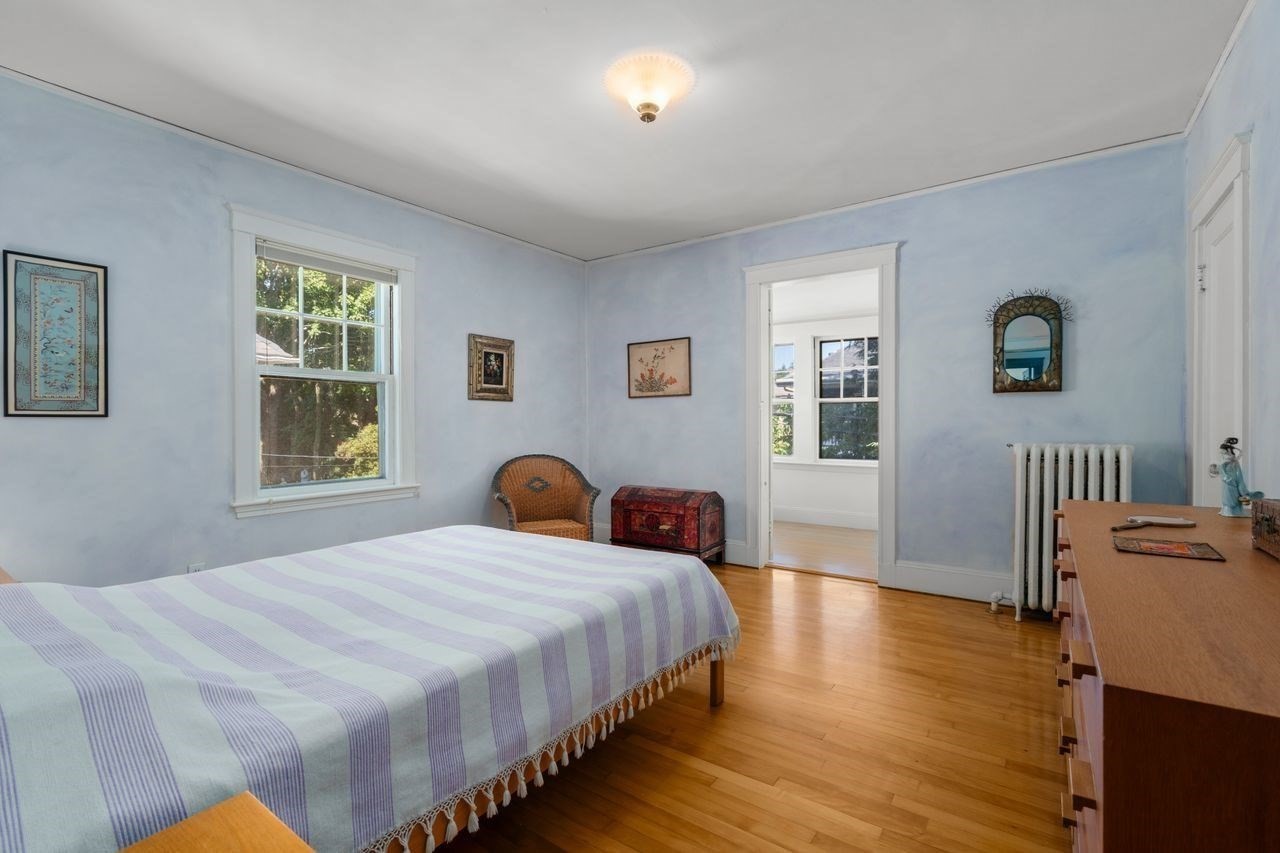 Bedroom with pale blue walls, a striped bedspread, hardwood floors, and windows overlooking greenery.