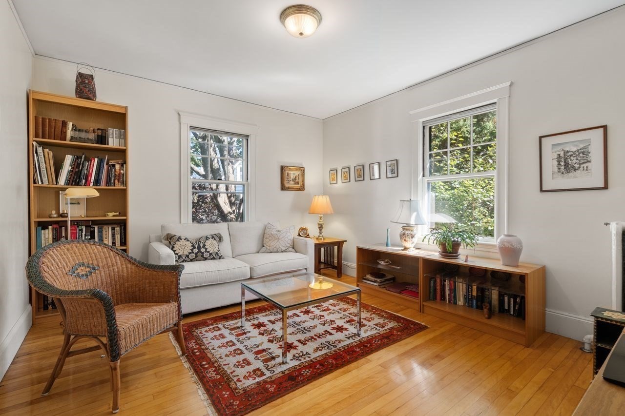 Living room with a white sofa, wicker armchair, wooden bookcase, and large windows overlooking leafy trees.