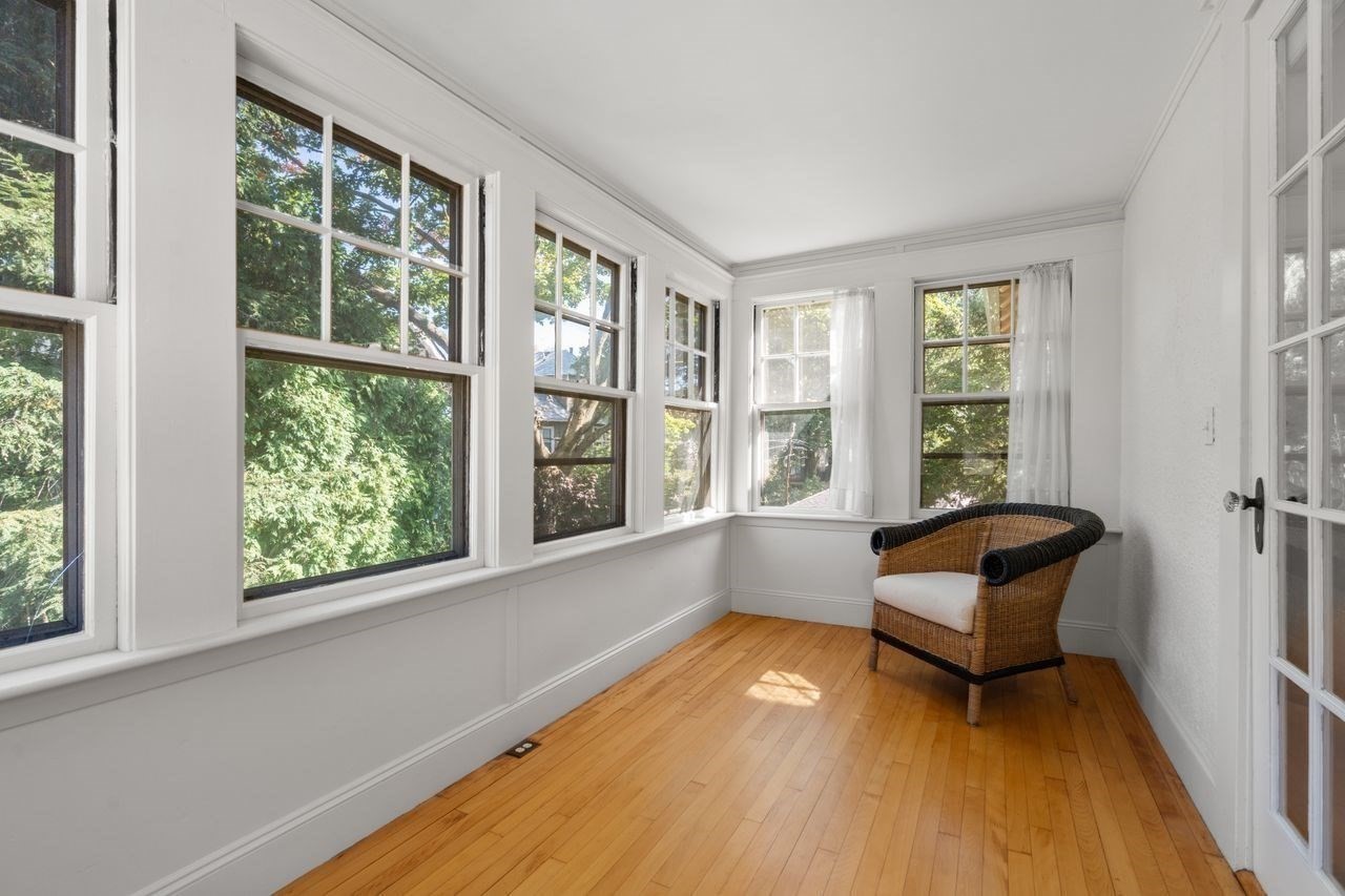 Bright sunroom with large leafy-view windows, hardwood floors, and a wicker chair.