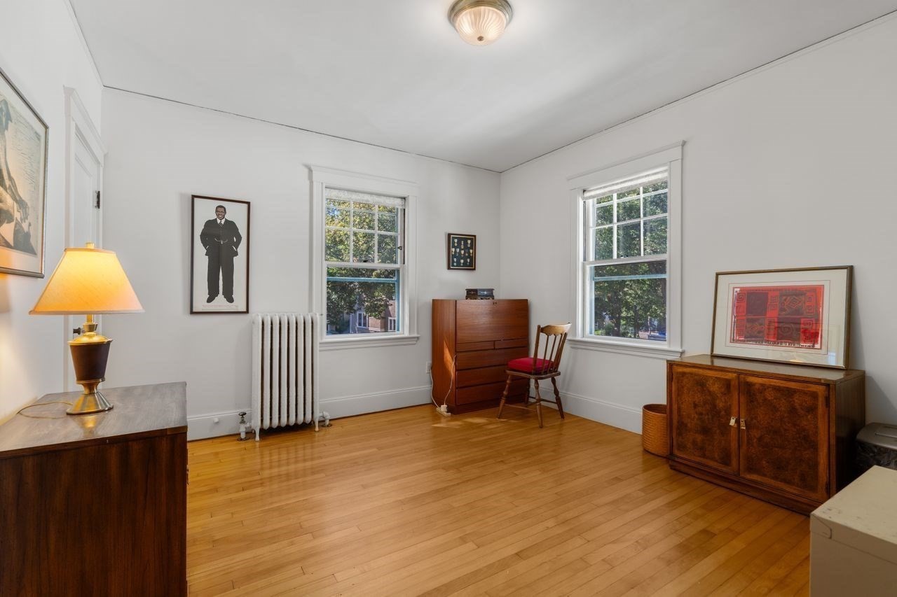 Furnished room with hardwood floors, two tall windows, a radiator, and wooden cabinets.