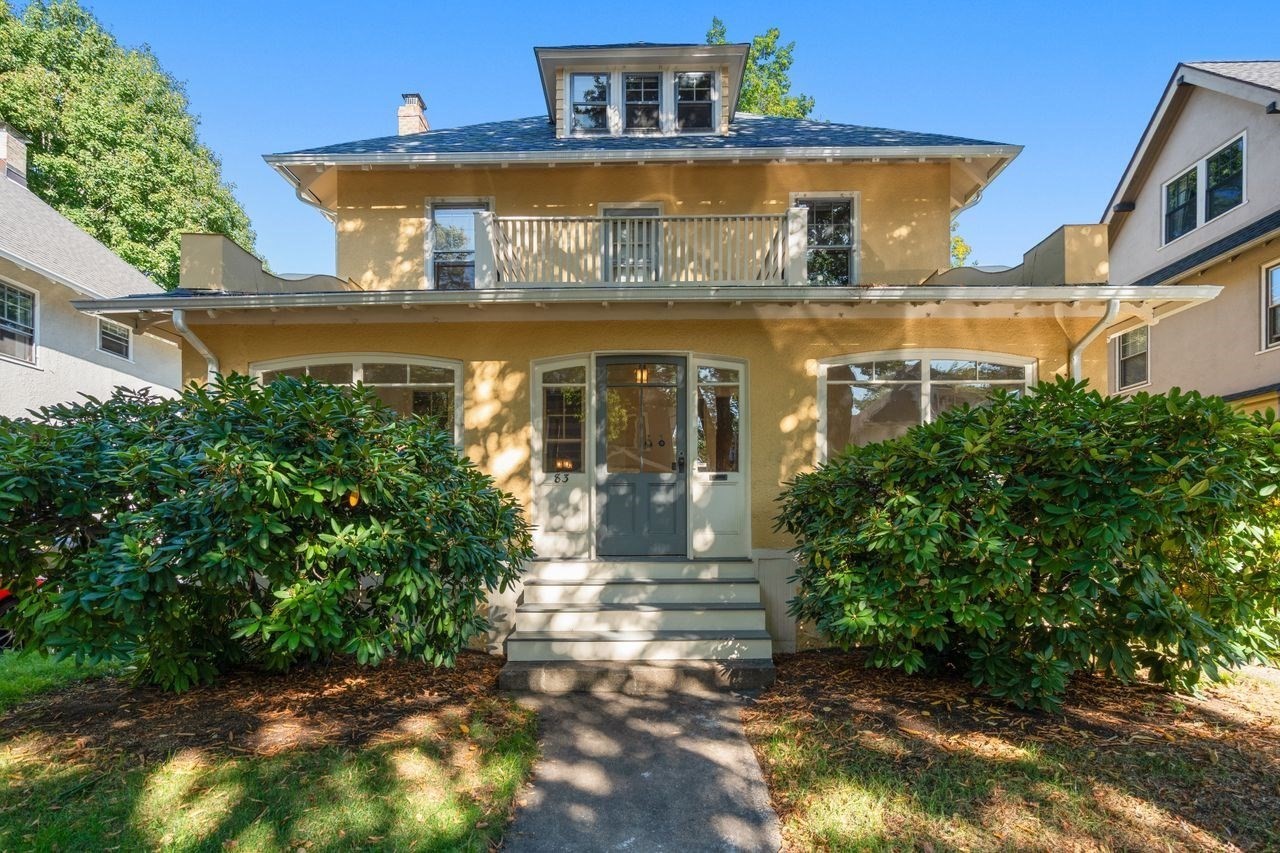 Ochre two-story home with an upper balcony, front steps, and mature shrubs.