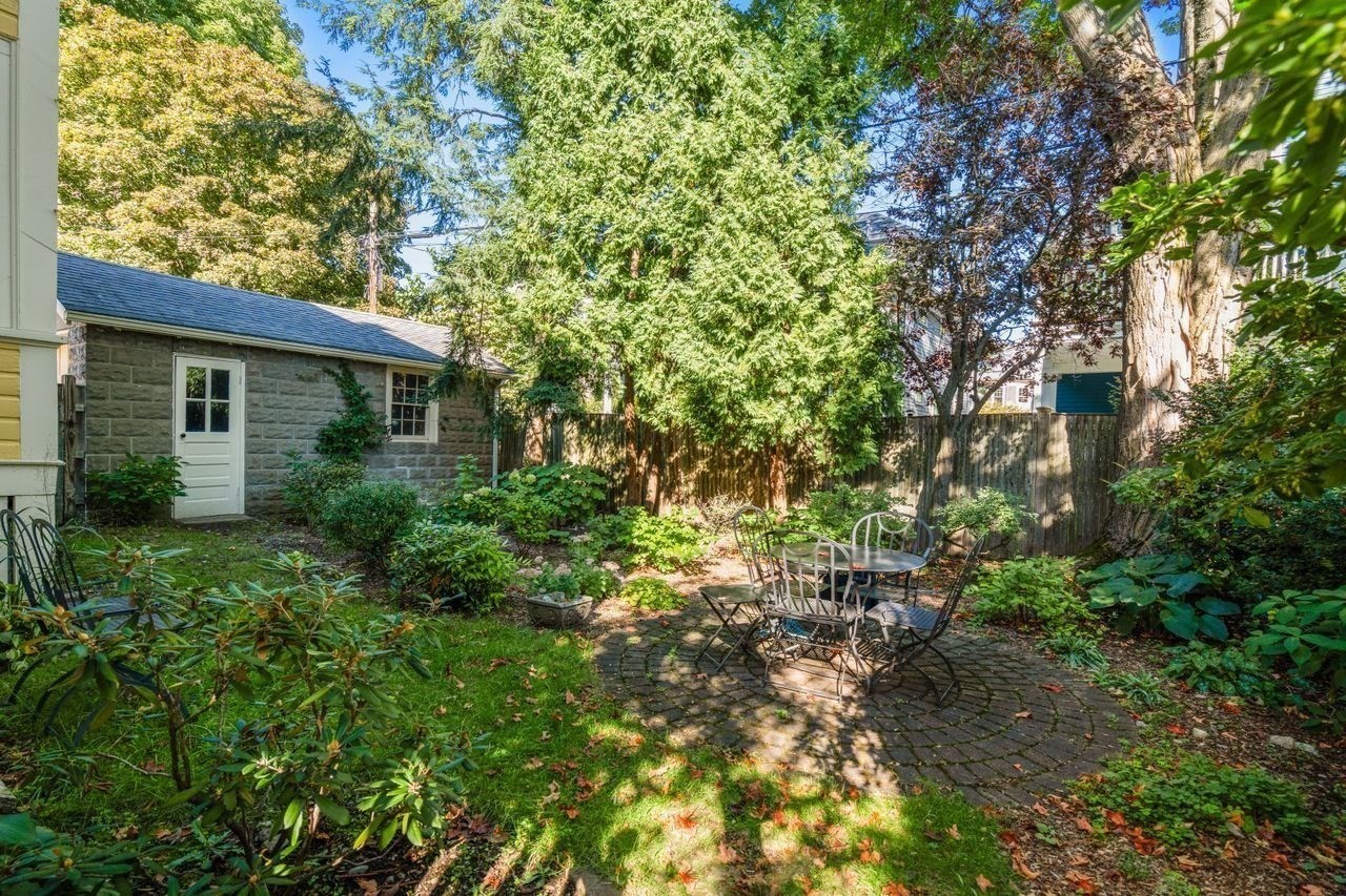 Backyard patio with a metal dining set, mature trees, and a small stone outbuilding.