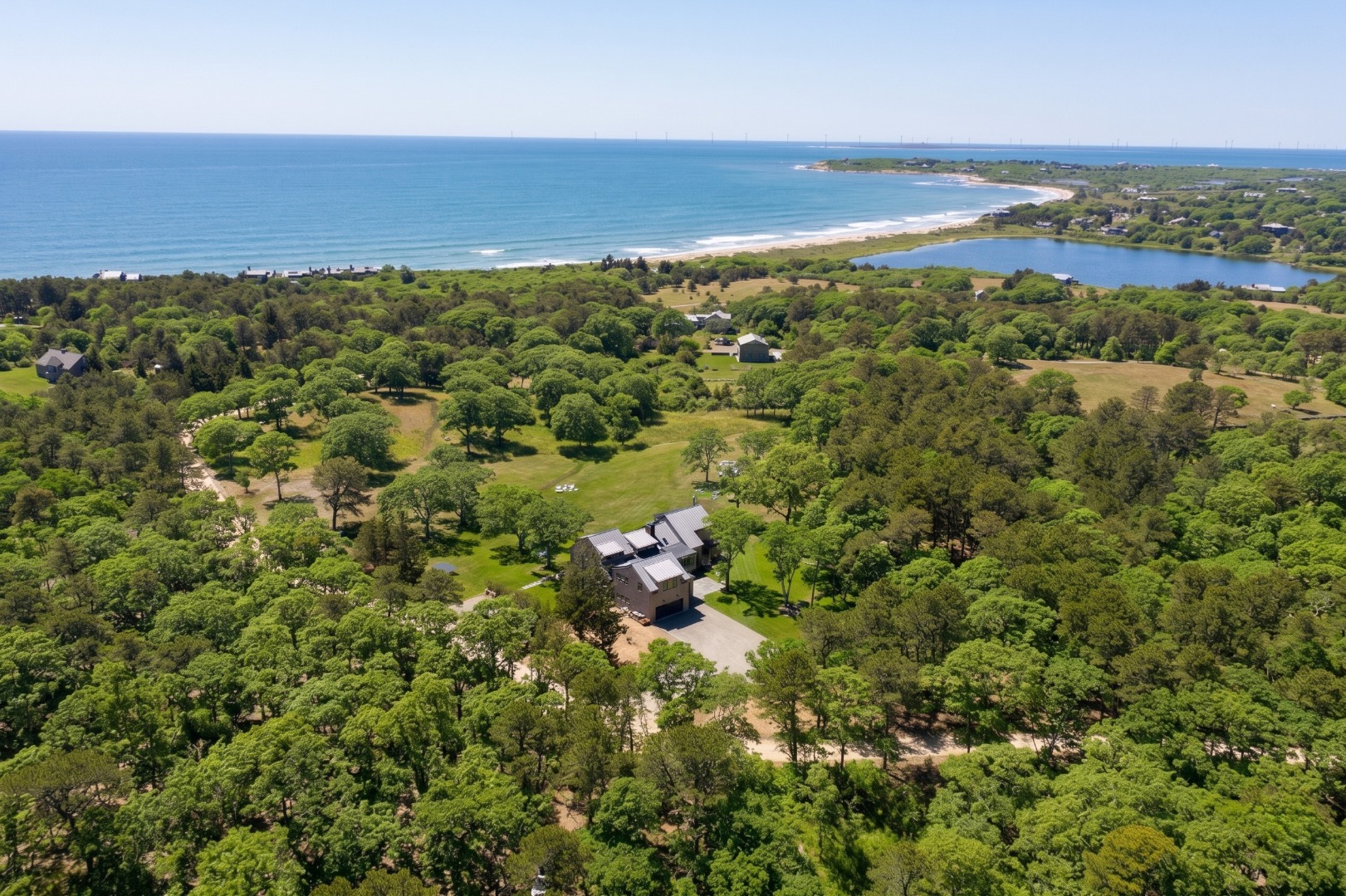 Aerial view of wooded coastal grounds, houses, a pond, and a curving beach beside the ocean.