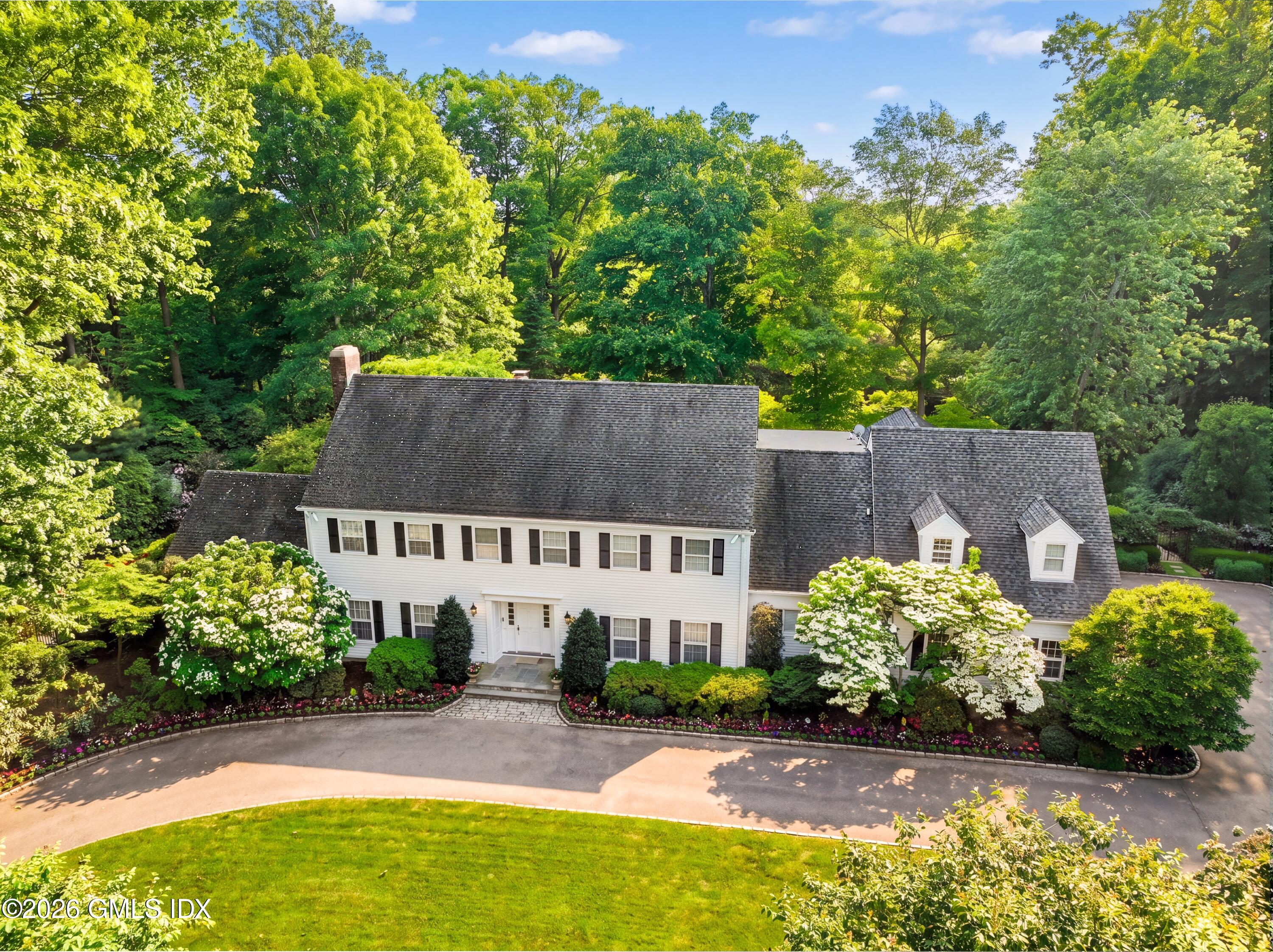 White two-story house with dark shutters, surrounded by mature trees and landscaped gardens.