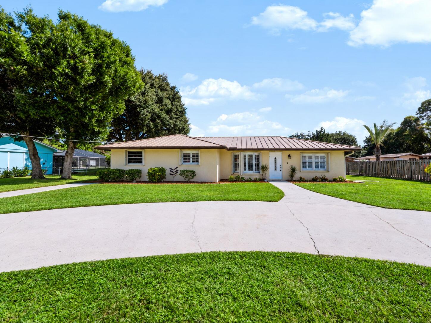 Single-story home with a brown metal roof, white shutters, and a curved driveway across a broad lawn.