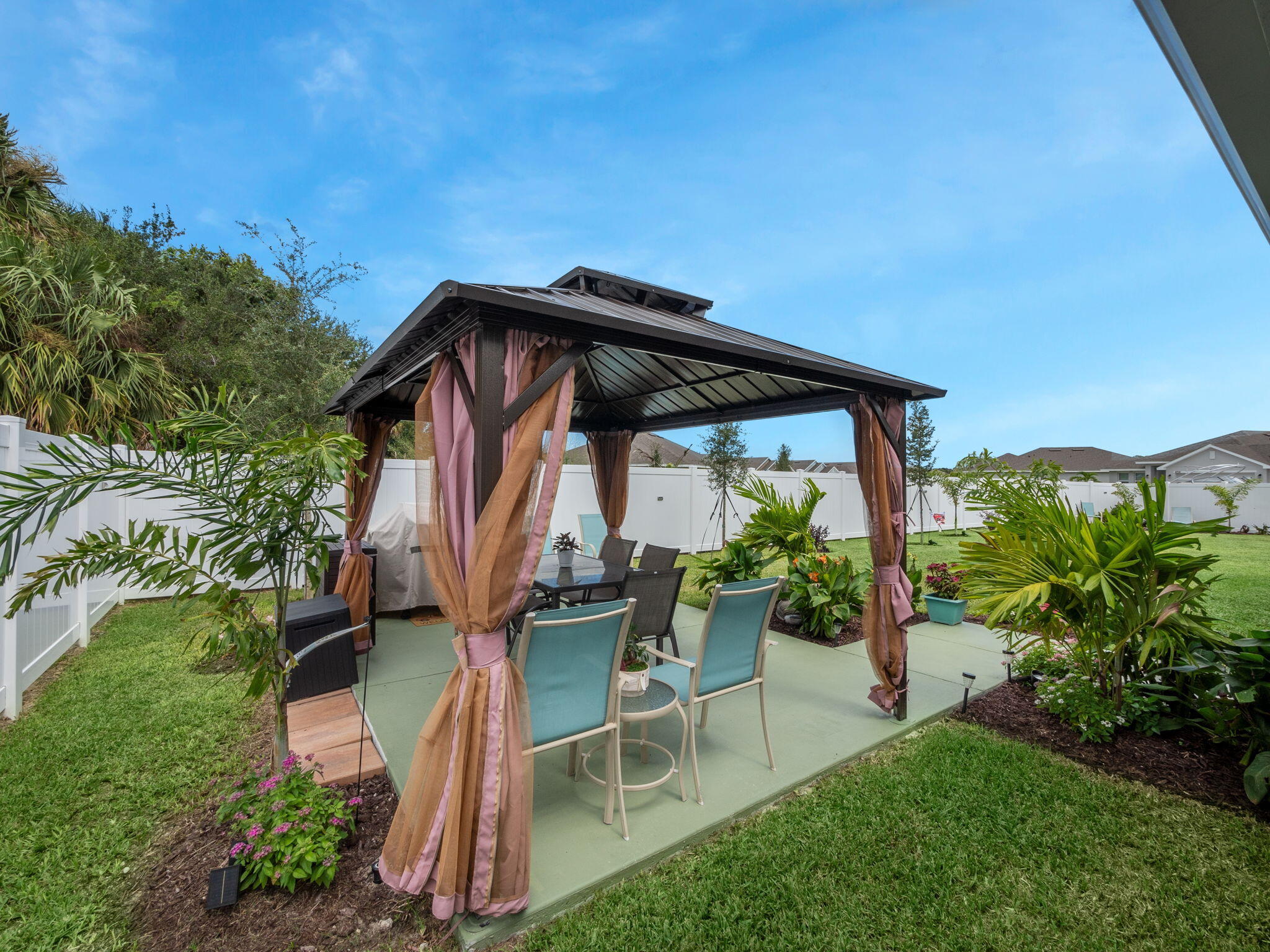 Backyard patio beneath a curtained pergola, furnished with outdoor dining chairs and surrounded by tropical landscaping.