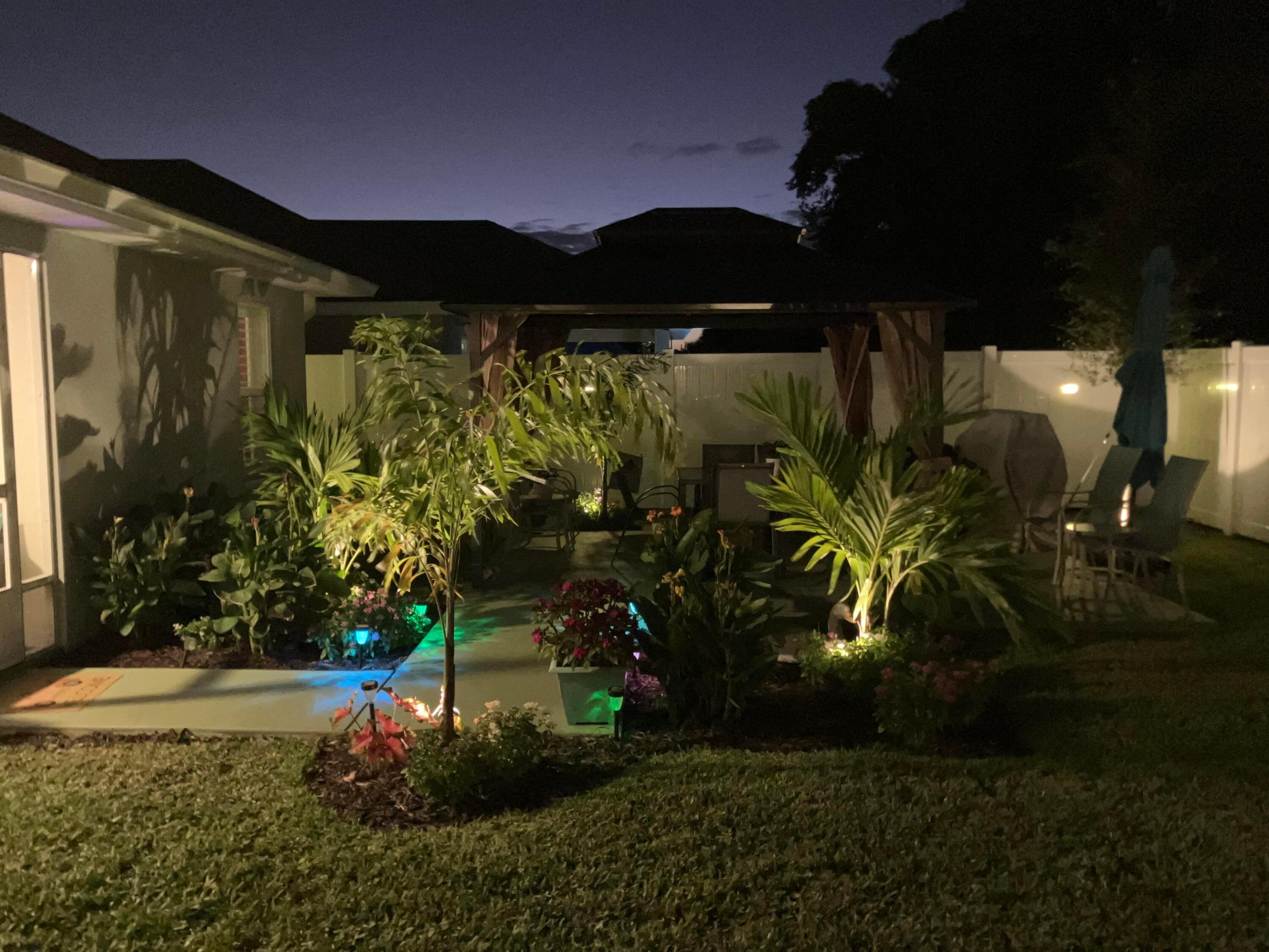 Landscaped backyard patio at night with a covered seating area and colorful lighting among tropical plants.