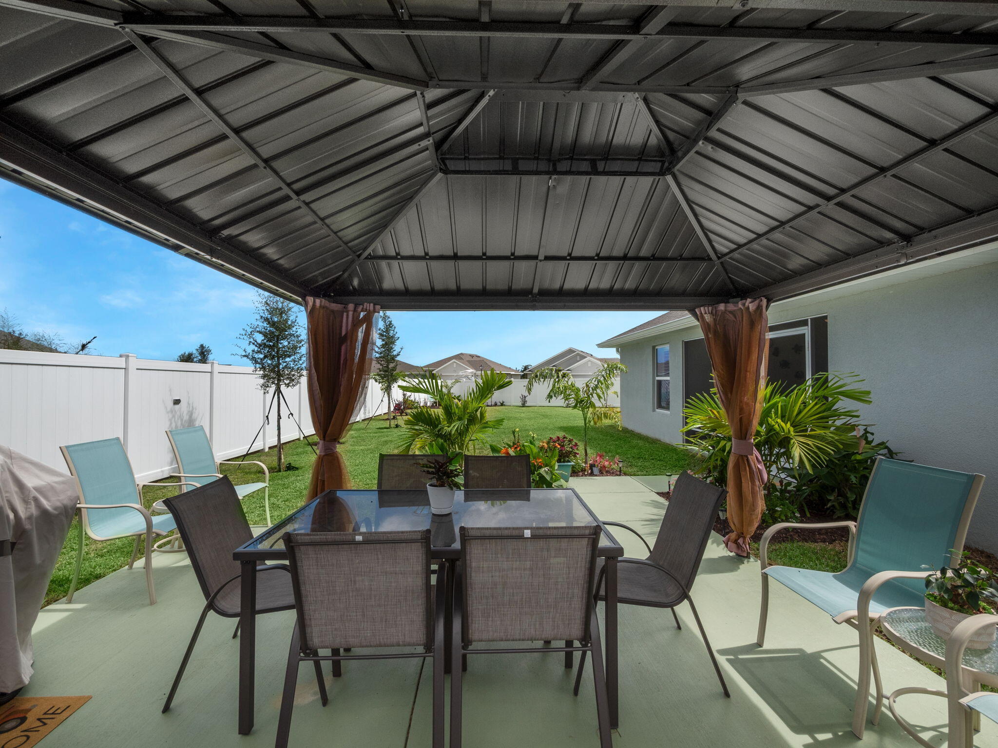 Covered backyard patio with a metal roof, outdoor dining set, tropical plants, and fenced lawn.