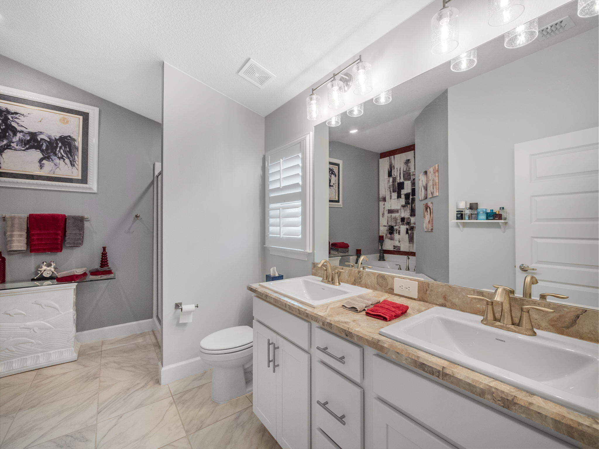 Bathroom with a double vanity, large mirror, plantation shutters, and pale tiled floors.