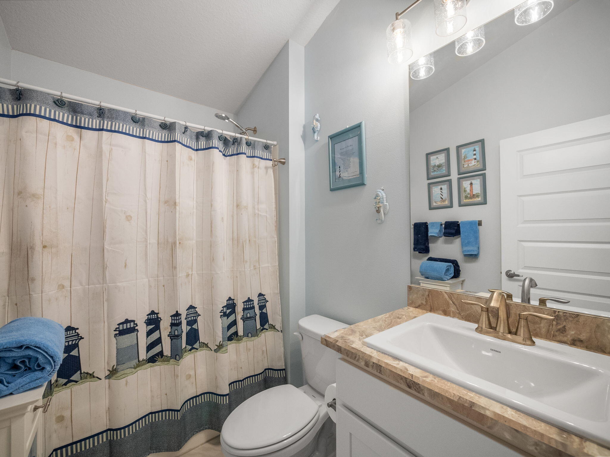 Bathroom with a lighthouse-patterned shower curtain, white vanity, bronze faucet, and blue towels.