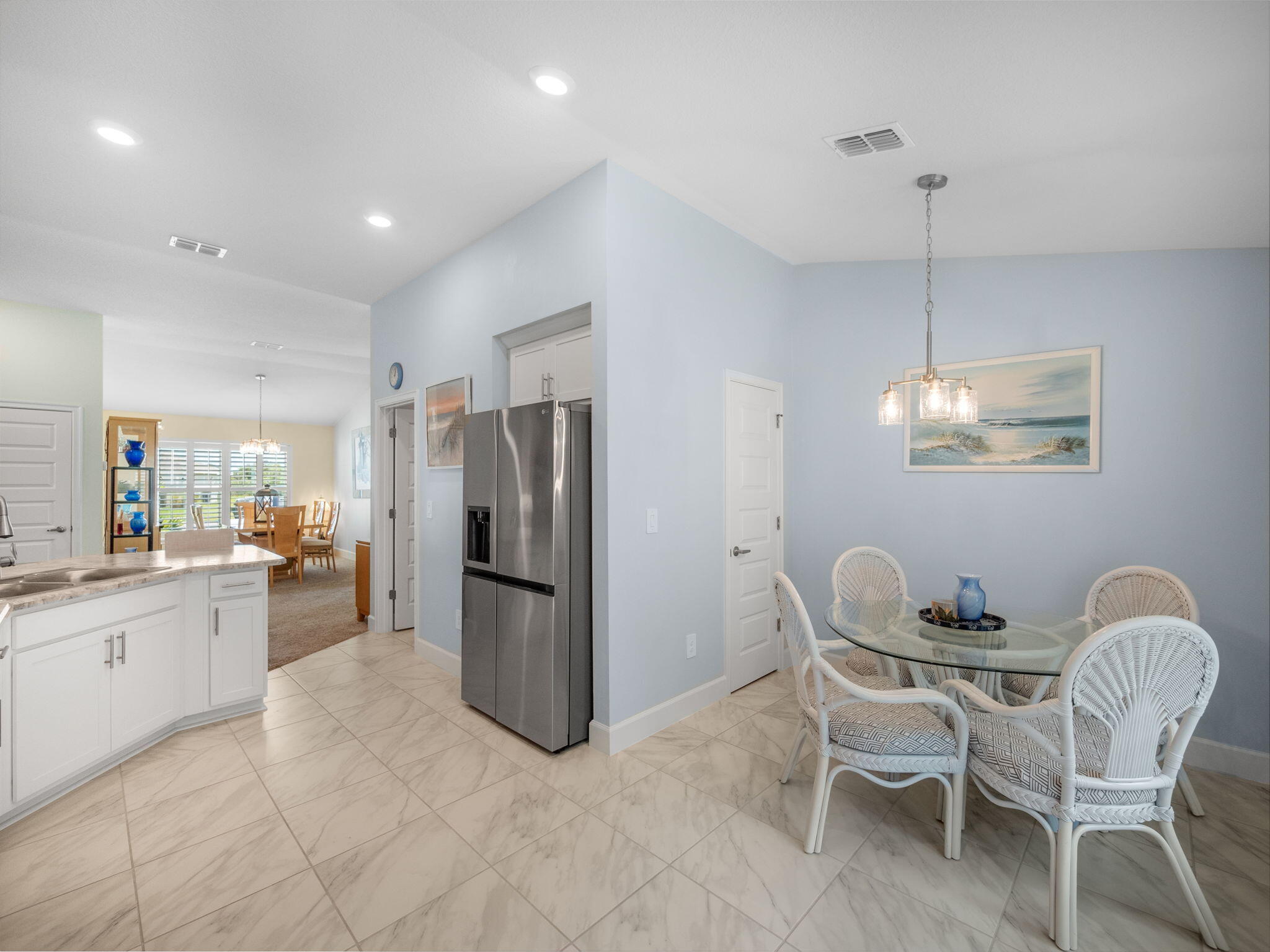 Open-plan kitchen and dining area with stainless-steel refrigerator and white woven chairs around a glass table.