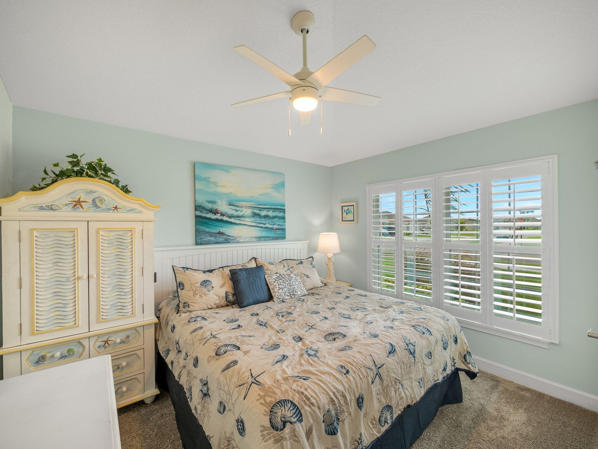 Bedroom with seashell-patterned bedding, a coastal armoire, and white plantation shutters.
