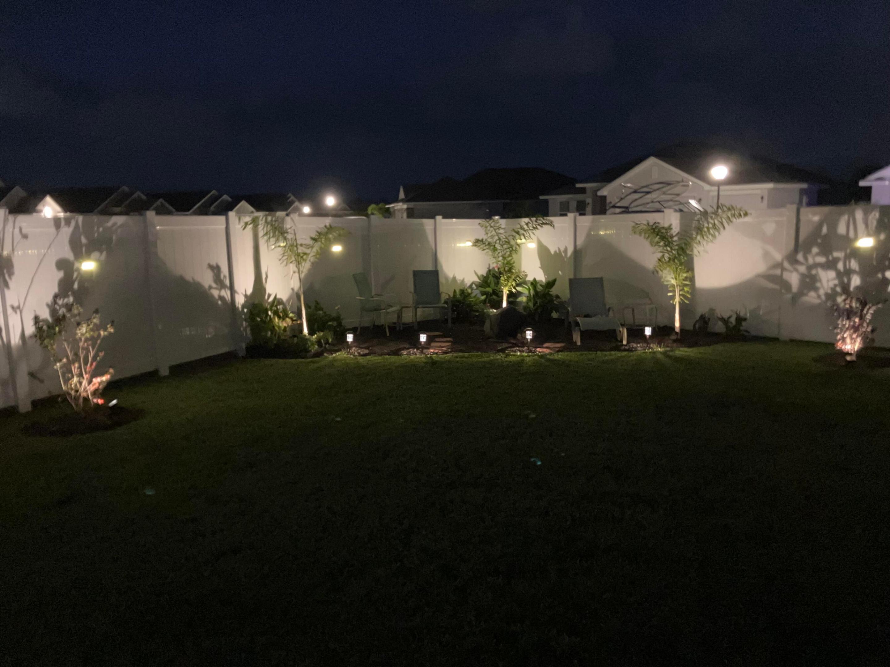 Backyard lawn with white privacy fence, landscaped seating area, and glowing garden lights at night.