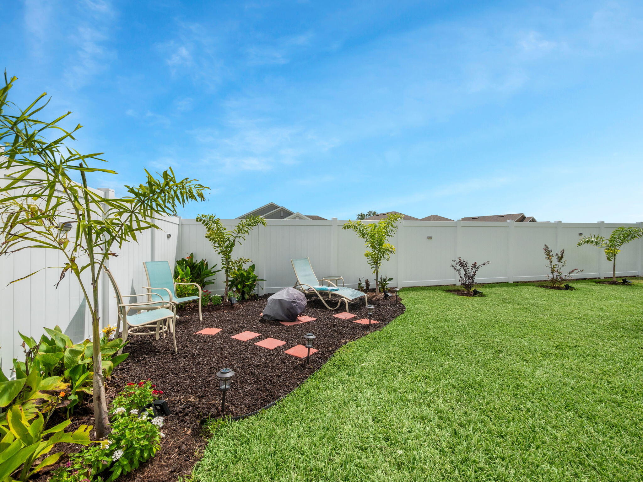 Backyard lawn bordered by tropical planting beds, lounge chairs, and a white privacy fence.