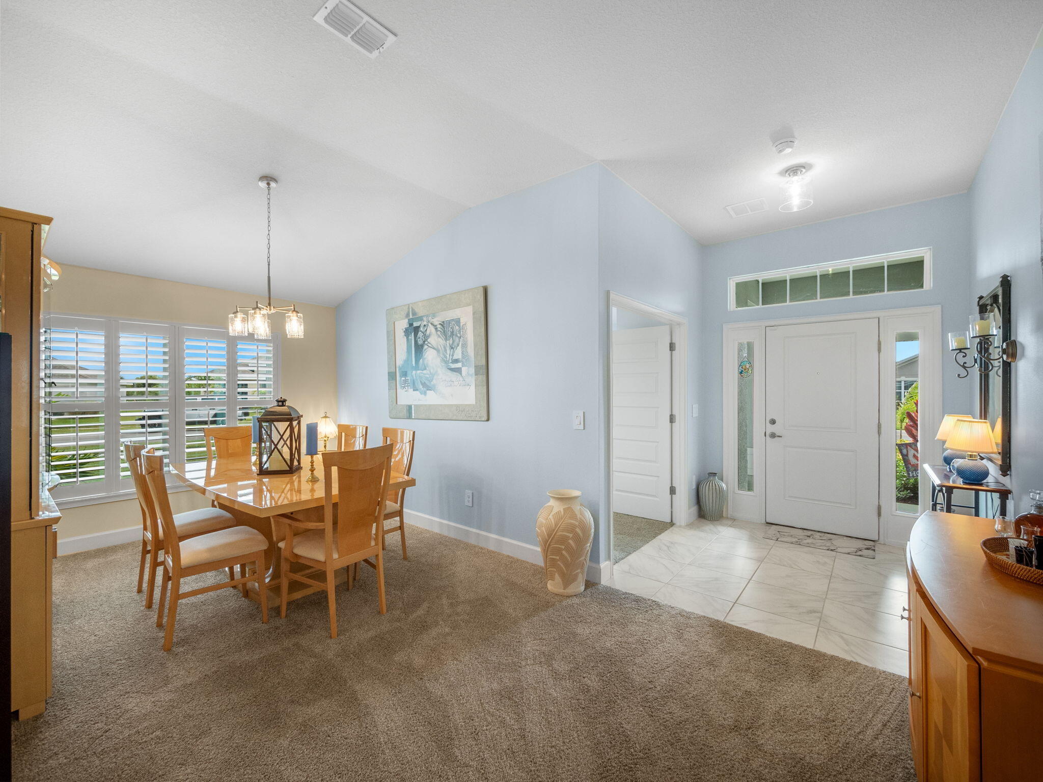 Dining area with wooden table and chairs beside shuttered windows, opening onto a tiled entryway.