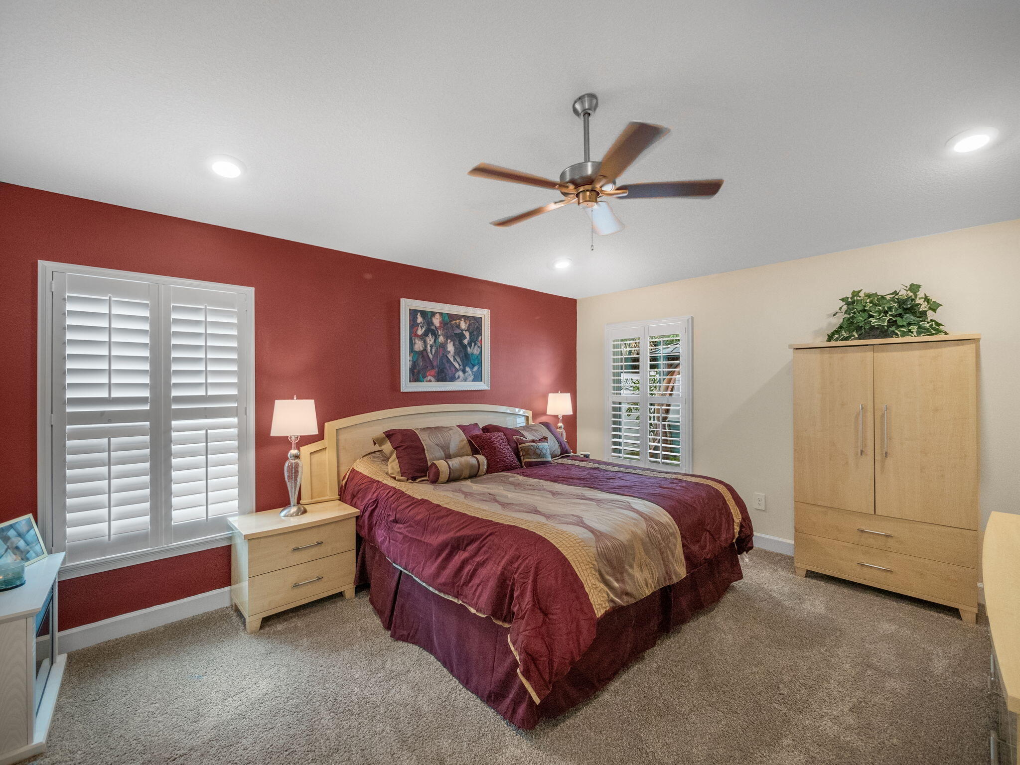 Bedroom with burgundy bedding, light wood furniture, plantation shutters, and a red accent wall.