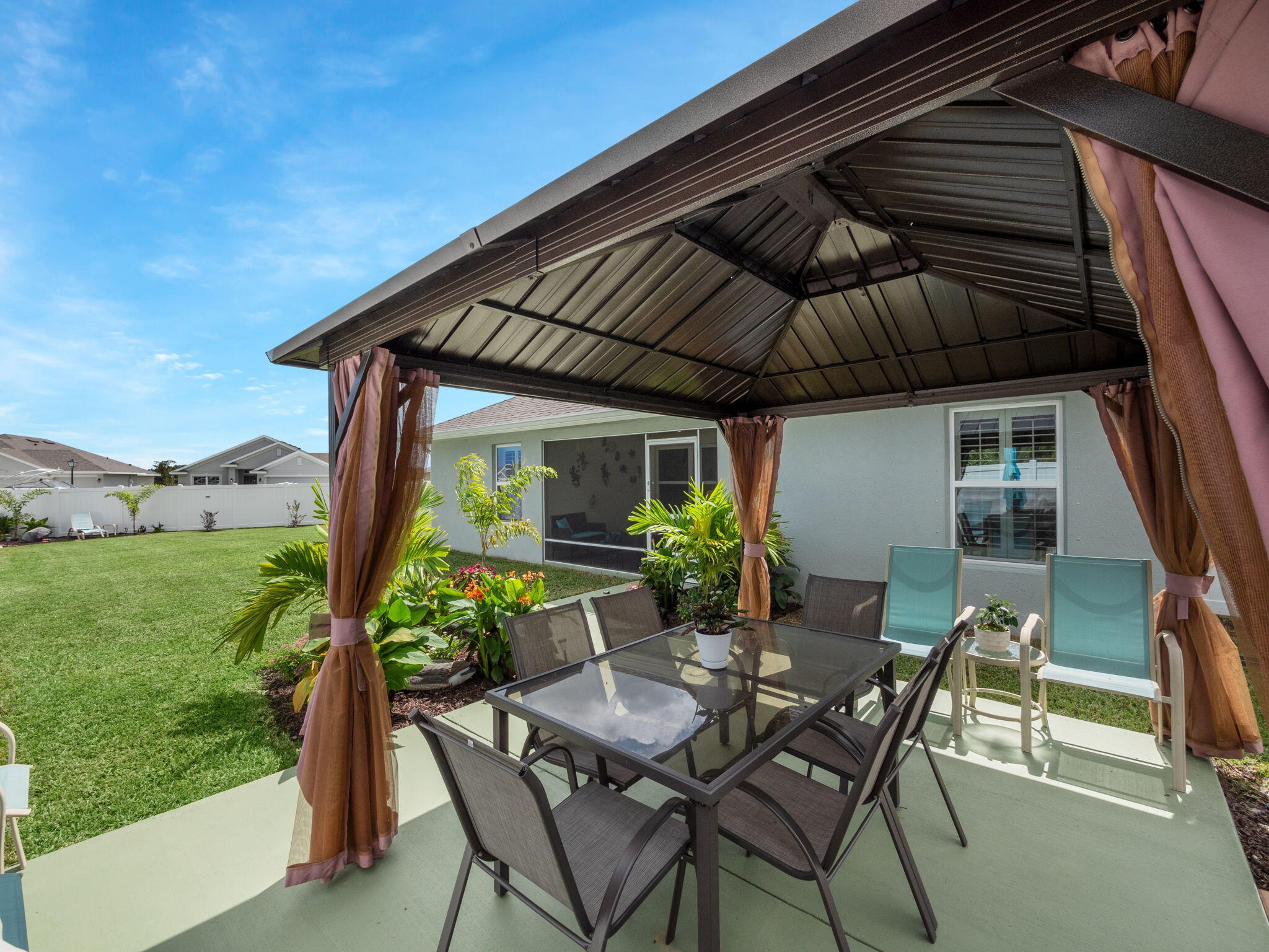 Backyard patio with a curtained metal gazebo, glass dining table, tropical plants, and lawn.