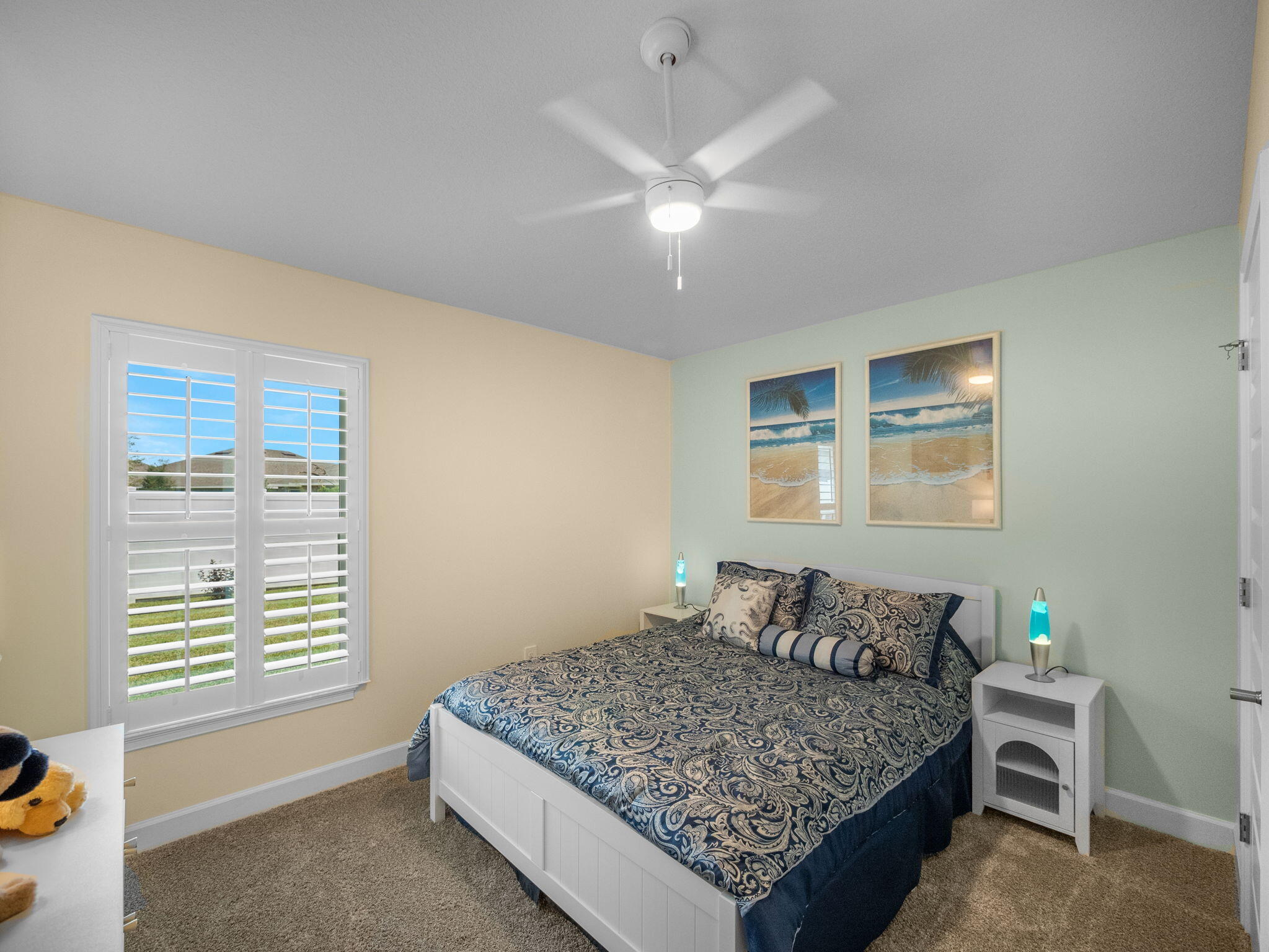 Furnished bedroom with blue patterned bedding, plantation shutters, and beach-themed wall art.