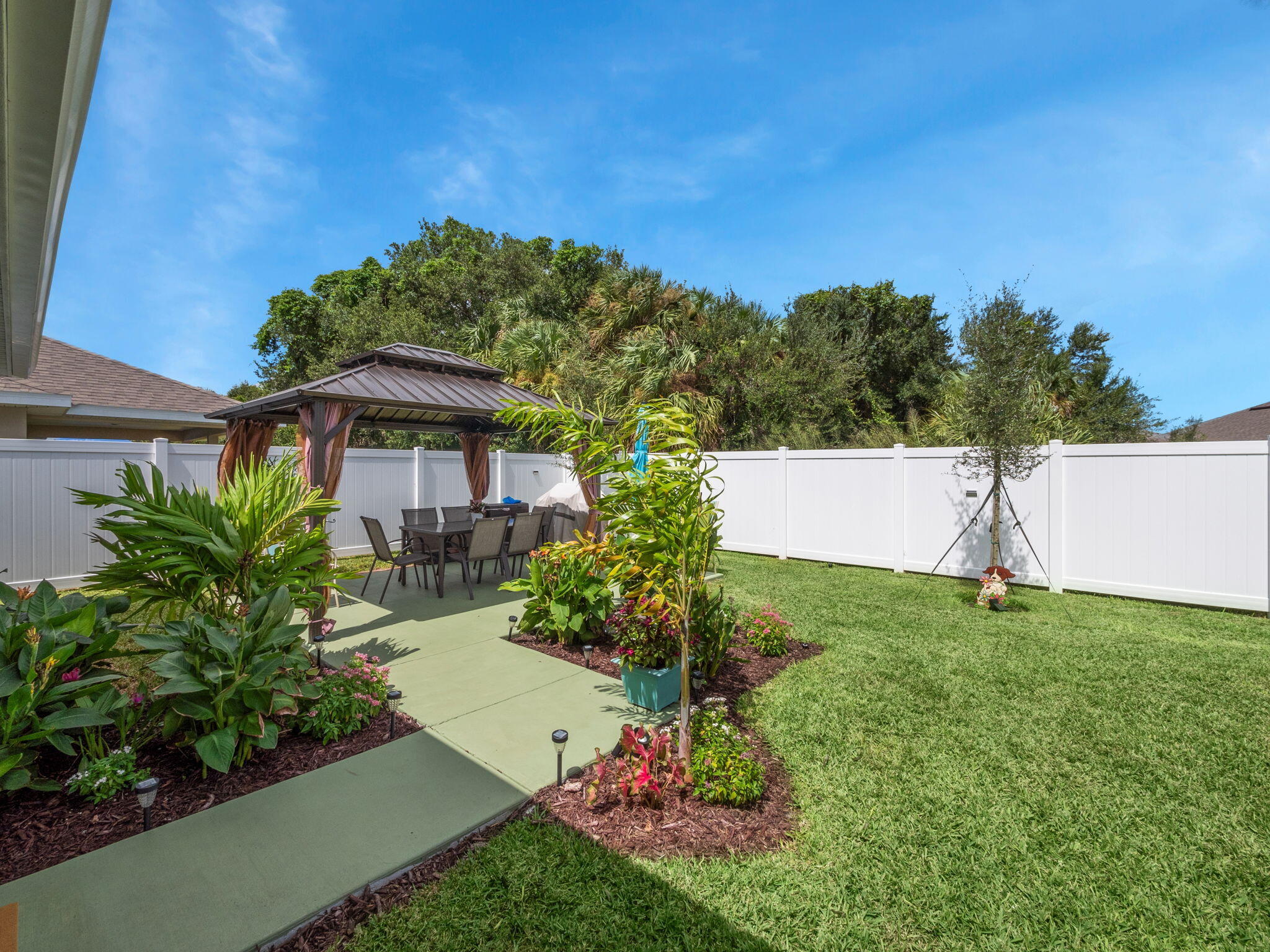 Landscaped backyard patio with a gazebo, outdoor dining set, tropical plants, and white privacy fence.