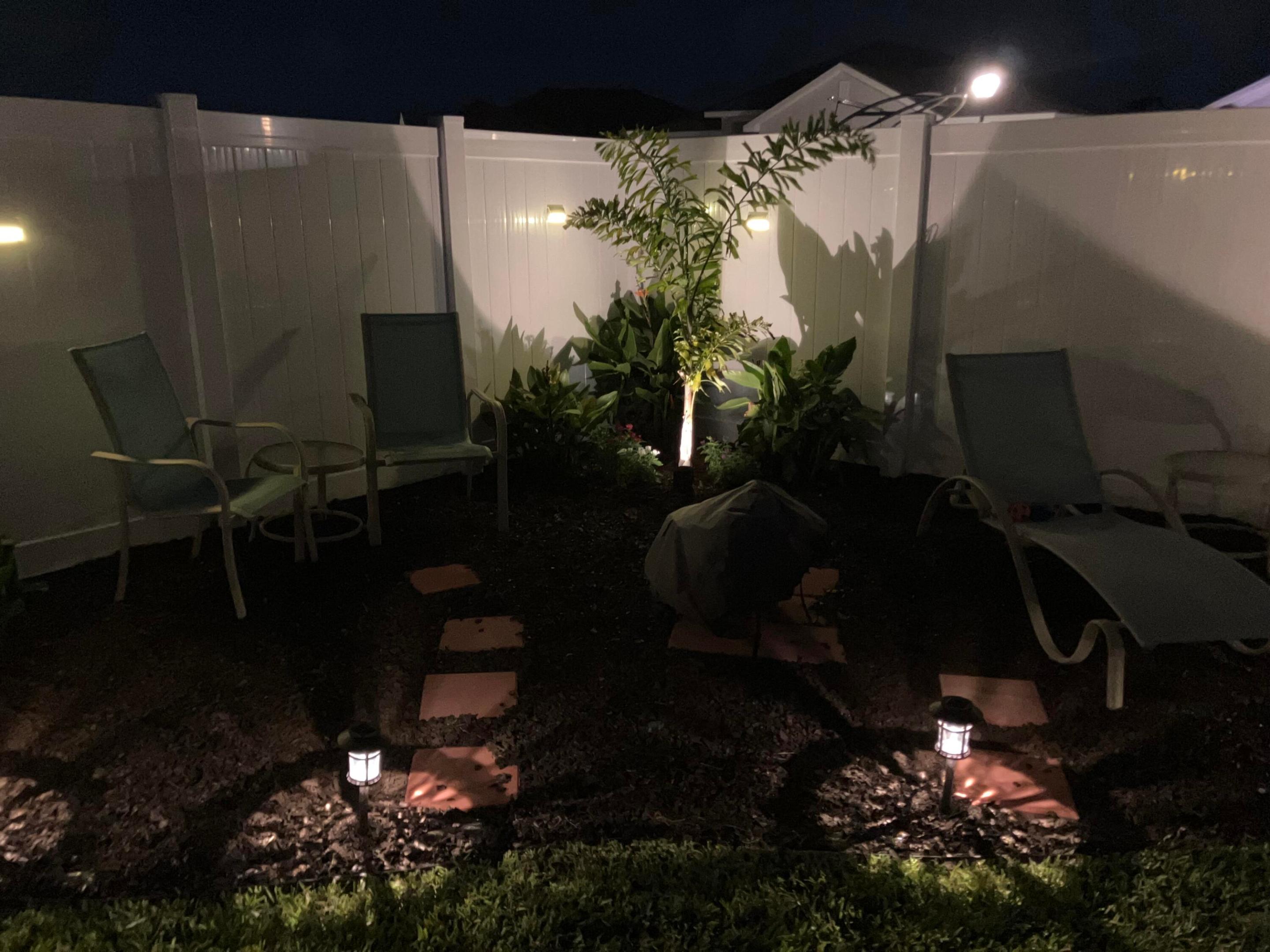 Fenced patio at night with lounge chairs, stepping stones, and illuminated landscaping.