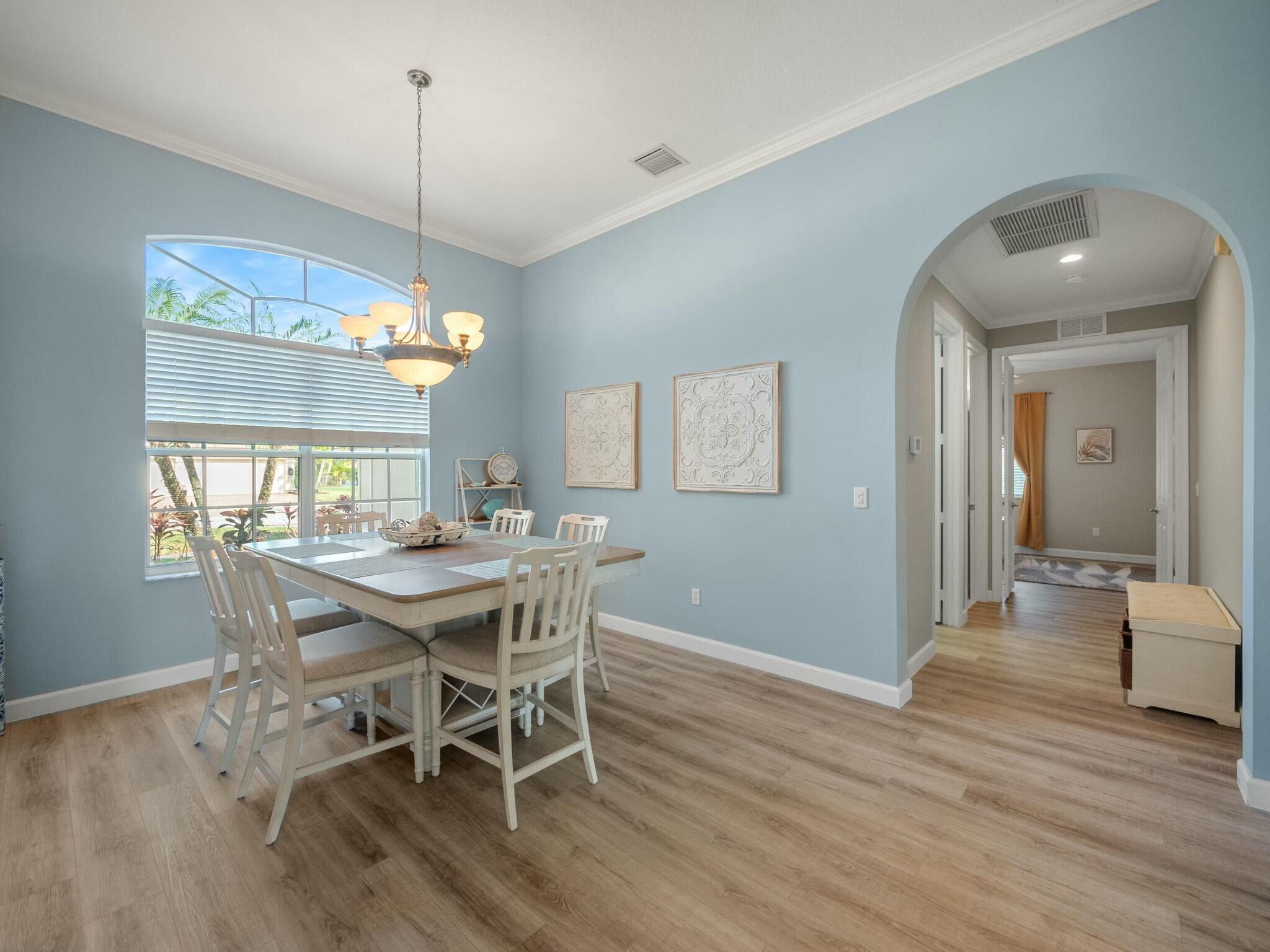 Dining room with a light wood table and chairs, chandelier, and large window overlooking tropical landscaping.