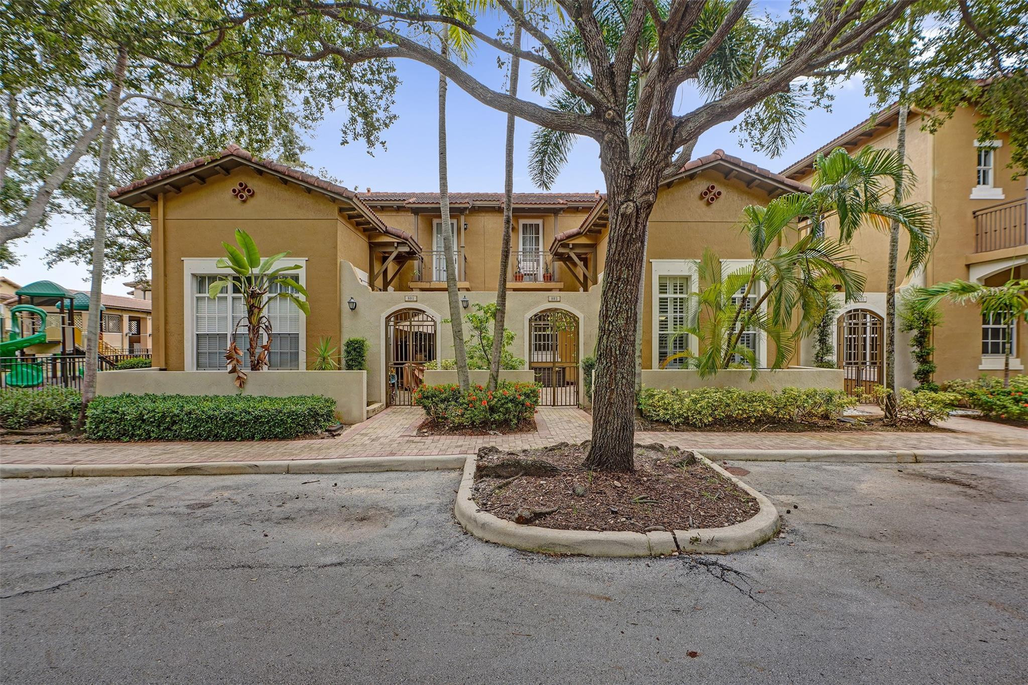 Tan two-story residential building with arched entrances, tropical landscaping, and a large shade tree in front.