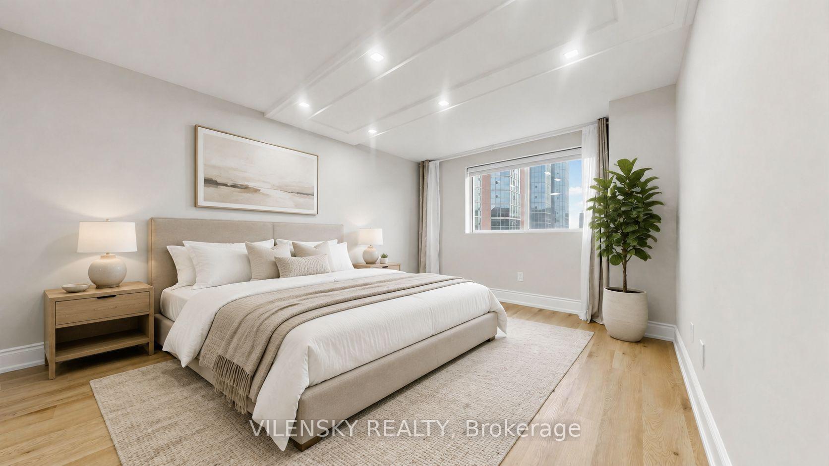 Staged bedroom with a beige upholstered bed, neutral rug, and window overlooking nearby high-rise buildings.