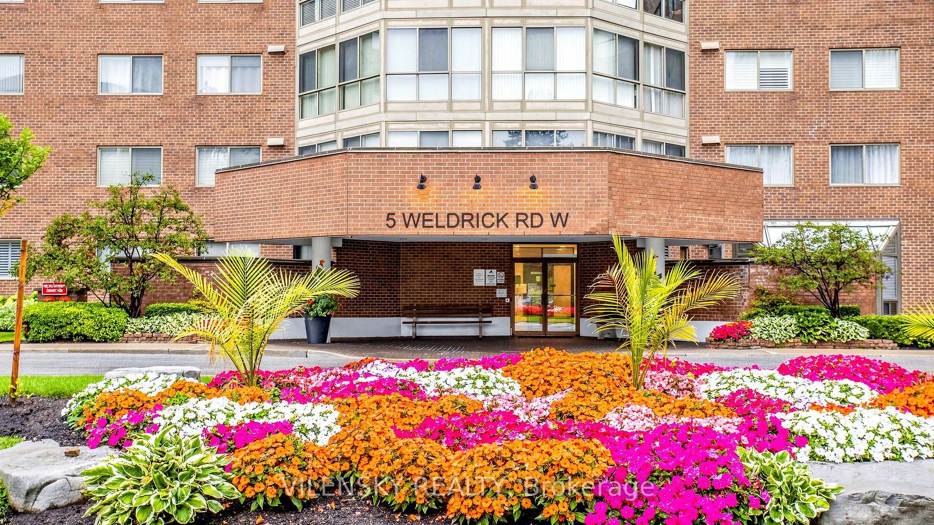 Brick condominium entrance marked 5 Weldrick Rd W, framed by colorful flowerbeds.