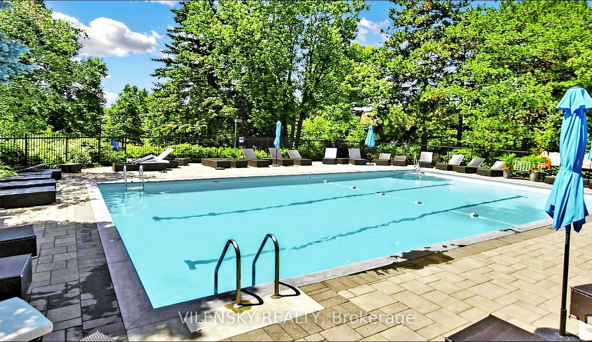 Outdoor swimming pool surrounded by trees, with lounge chairs and blue umbrellas on the deck.