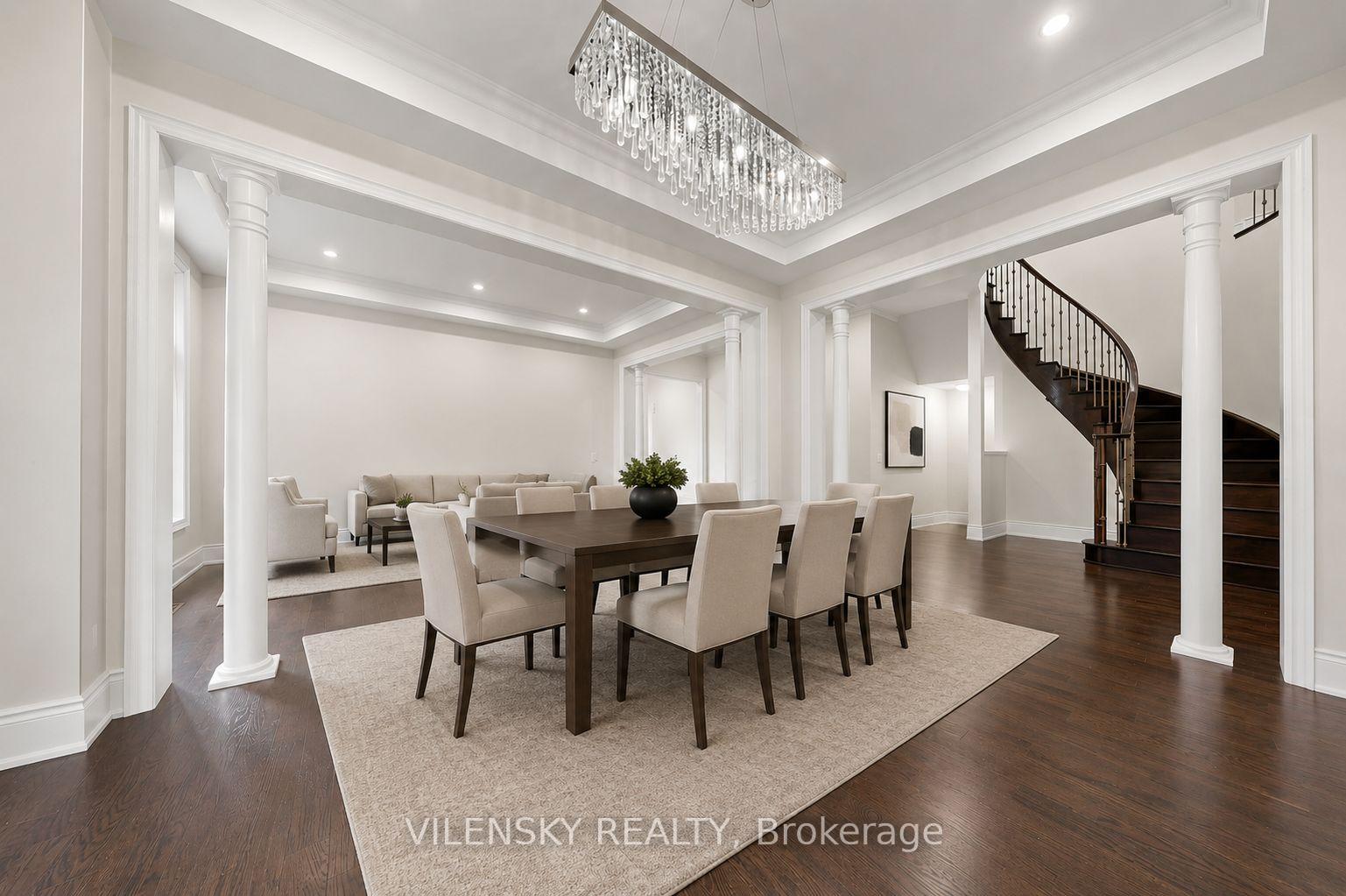 Dining room with a dark wood table, crystal chandelier, white columns, and curved staircase.