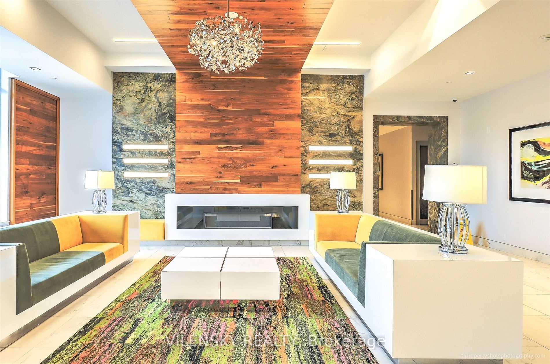 Condominium lobby lounge with a sculptural chandelier, colorful seating, patterned rug, and wood-and-stone fireplace wall.