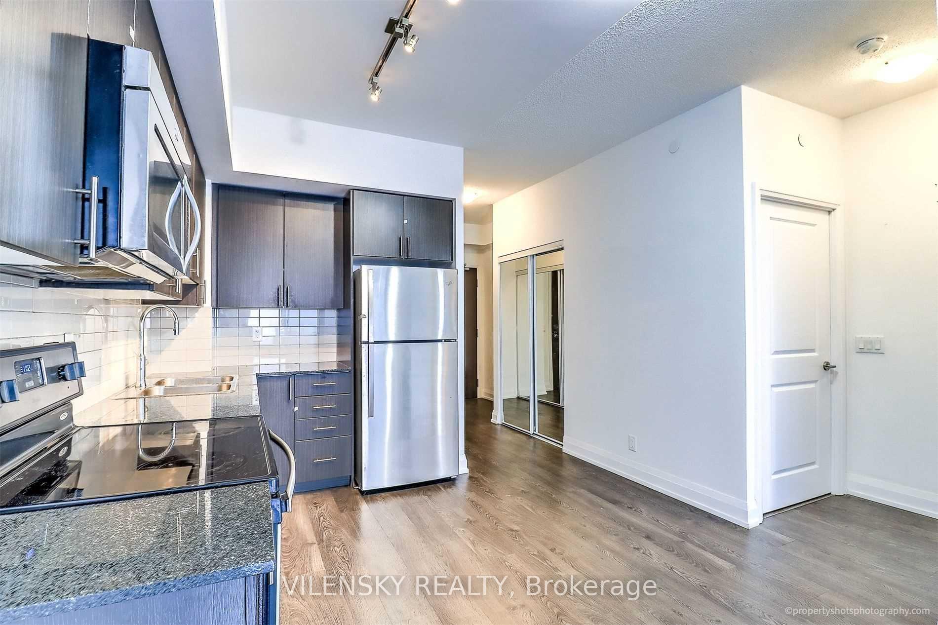 Modern condo kitchen with dark cabinets, granite counters, and stainless steel appliances opening to an empty living area.