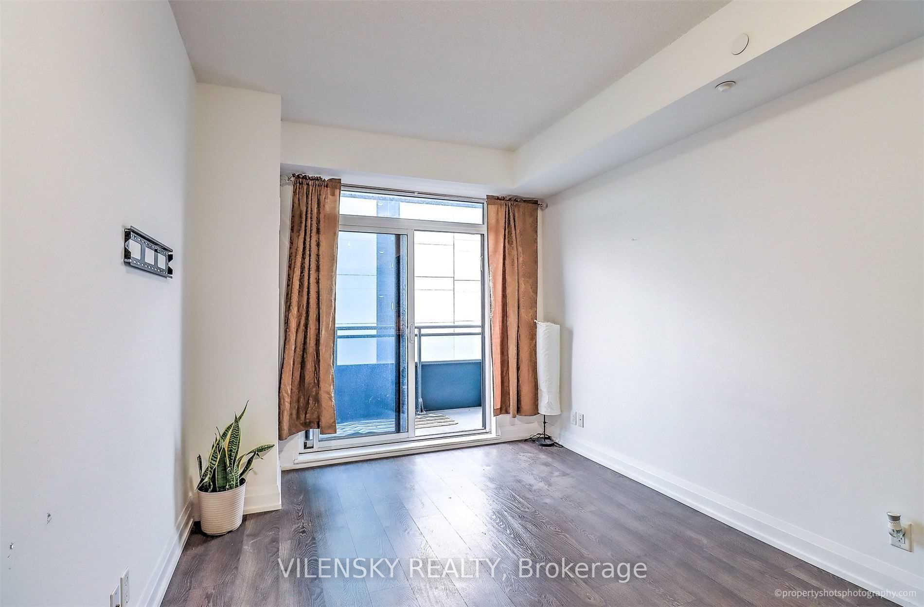 Mostly empty condo room with dark flooring, brown curtains, and a sliding glass door to the balcony.