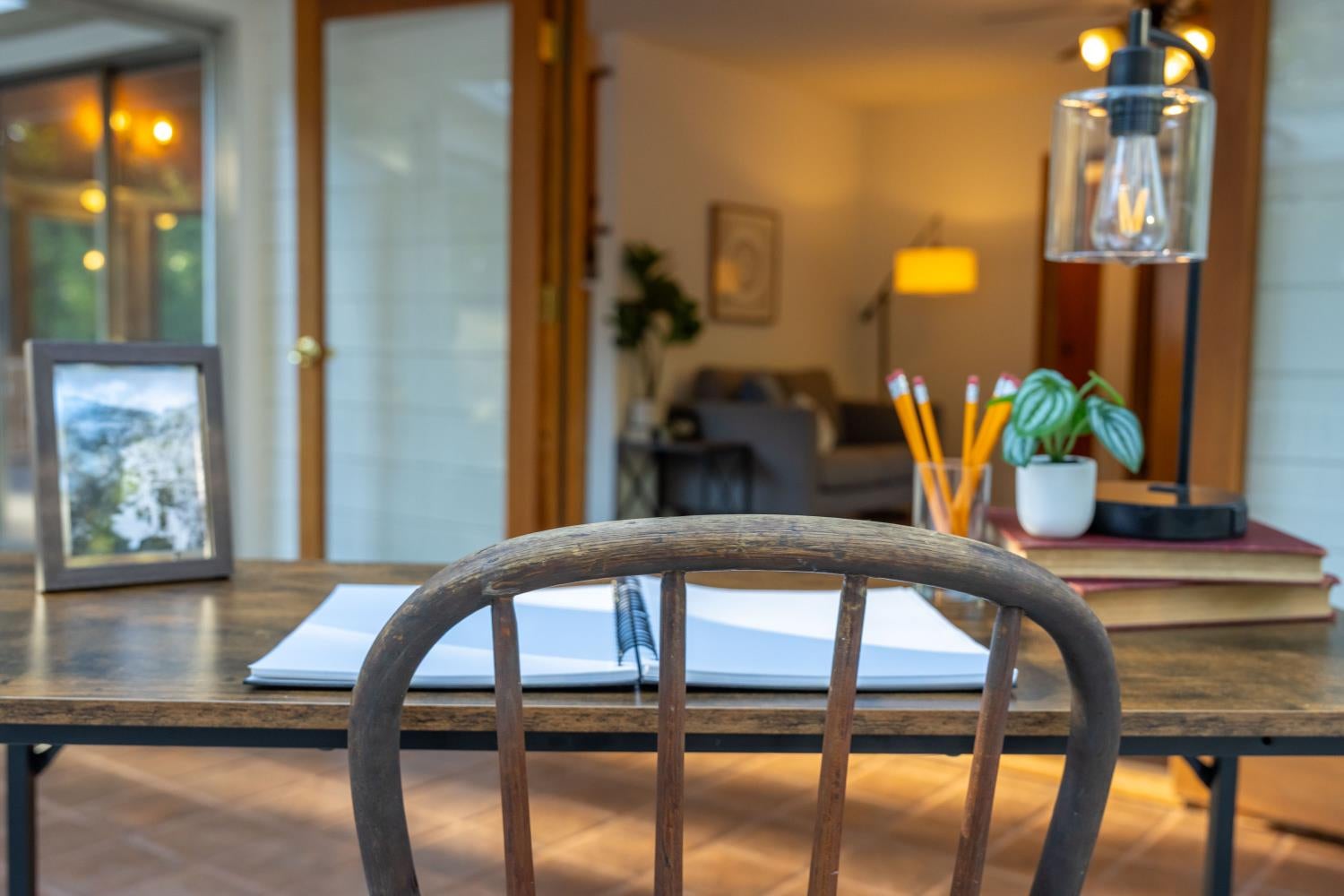 Wooden desk with an open notebook and pencils, with a sofa and lamp in the background.