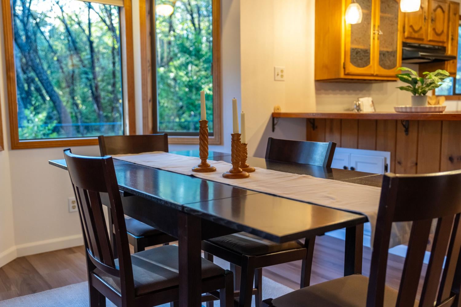 Dining room with a dark table beside large windows overlooking leafy trees and an adjacent wood-cabinet kitchen.