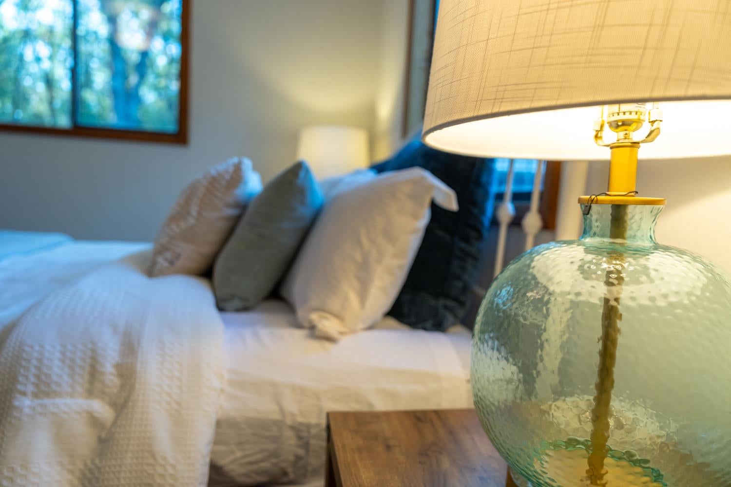 Bedroom with white bedding and layered pillows beside a glowing glass-base bedside lamp.