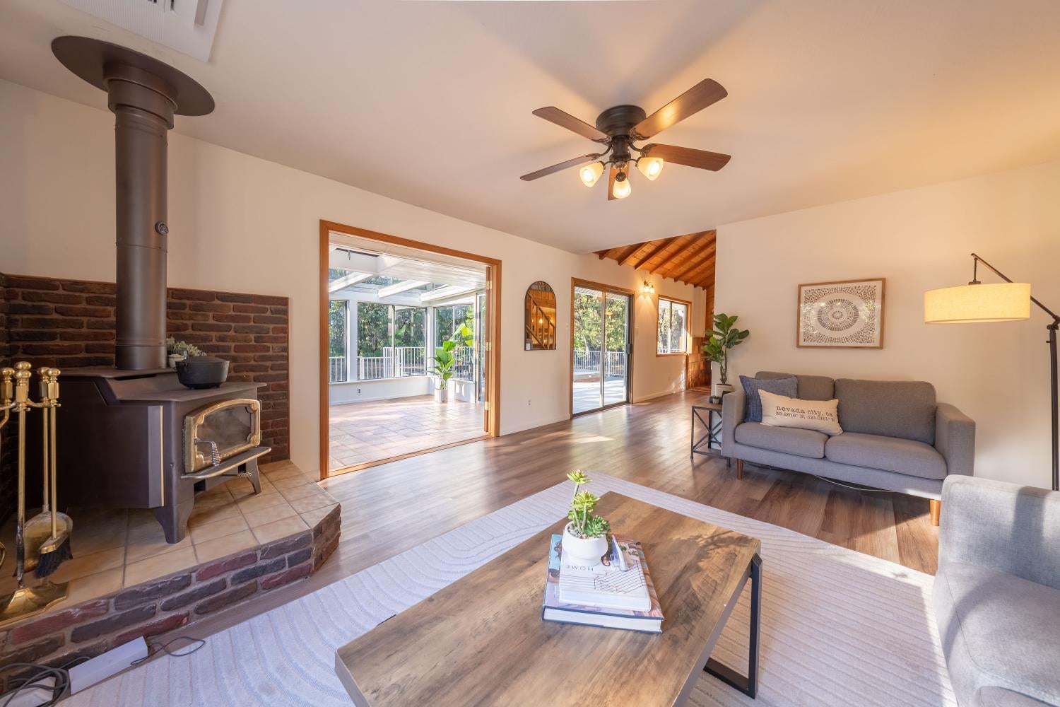 Living room with a brick-backed wood stove, gray sofa, and doorway opening to a bright sunroom.