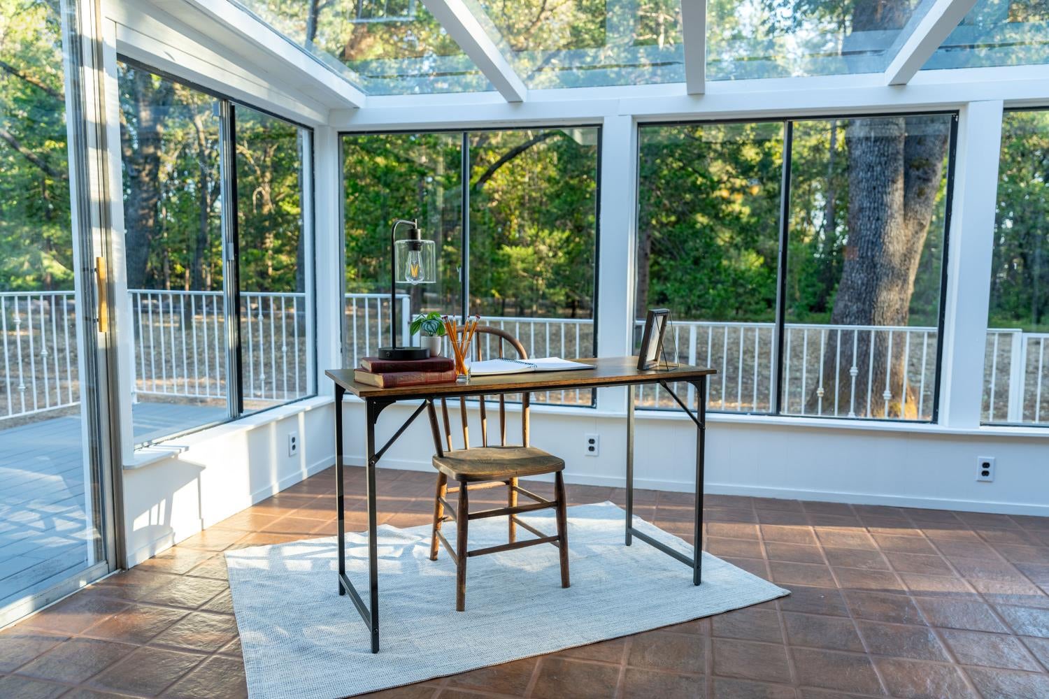 Sunroom office with a desk beneath a glass ceiling, overlooking trees and a deck.