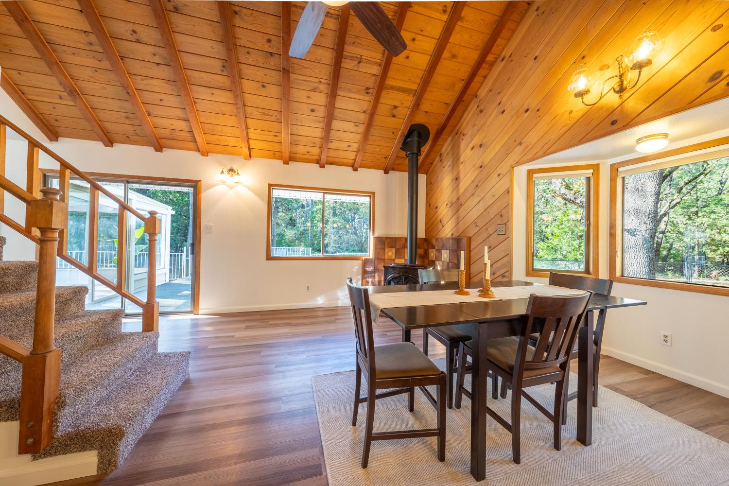 Dining area with vaulted wood ceiling, tiled fireplace, staircase, and windows overlooking trees.