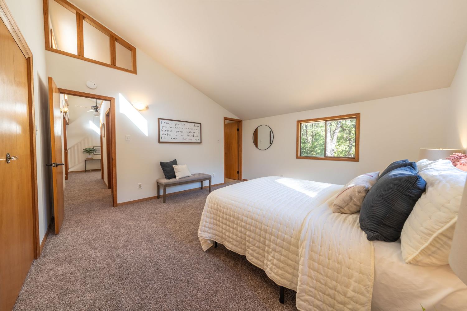 Furnished bedroom with a vaulted ceiling, high interior window, and views of trees.