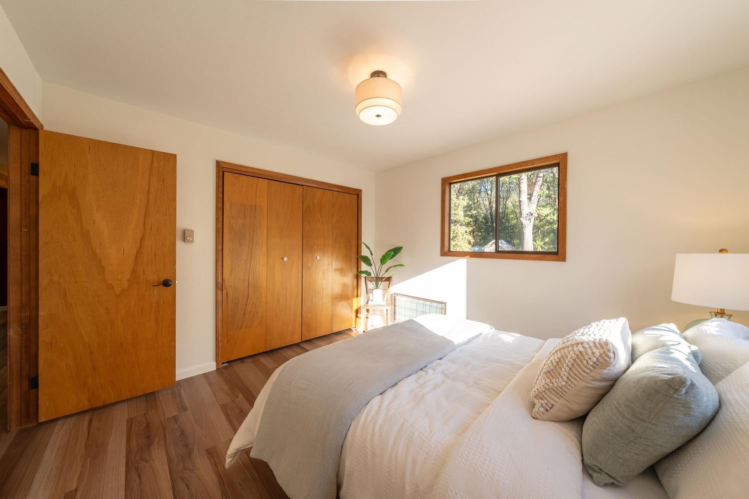 Bedroom with a white bed, warm wood closets, and a window overlooking trees.