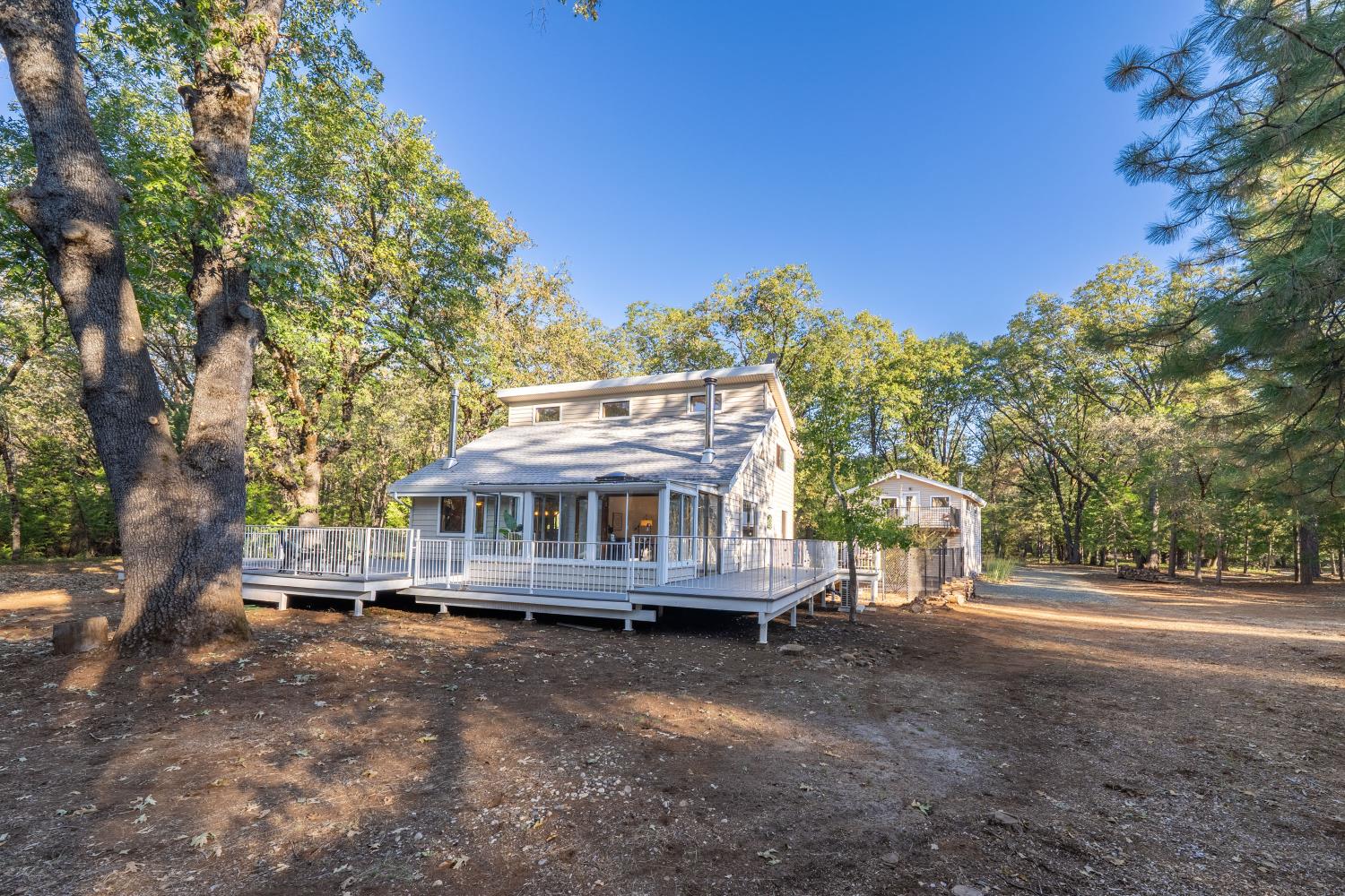 House with a wraparound deck among mature trees, with a smaller outbuilding beside it.