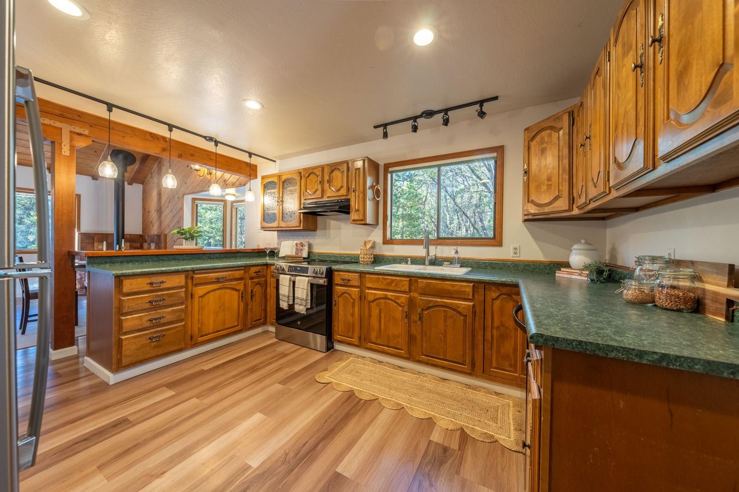 Kitchen with warm wood cabinets, green countertops, stainless appliances, and a window overlooking trees.