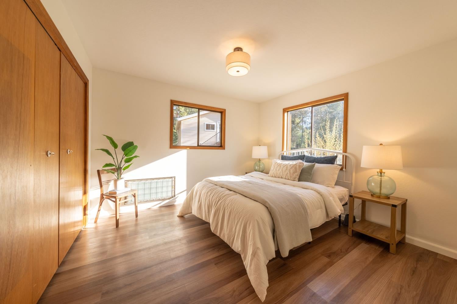 Staged bedroom with neutral bedding, warm wood floors, and two sunlit windows.